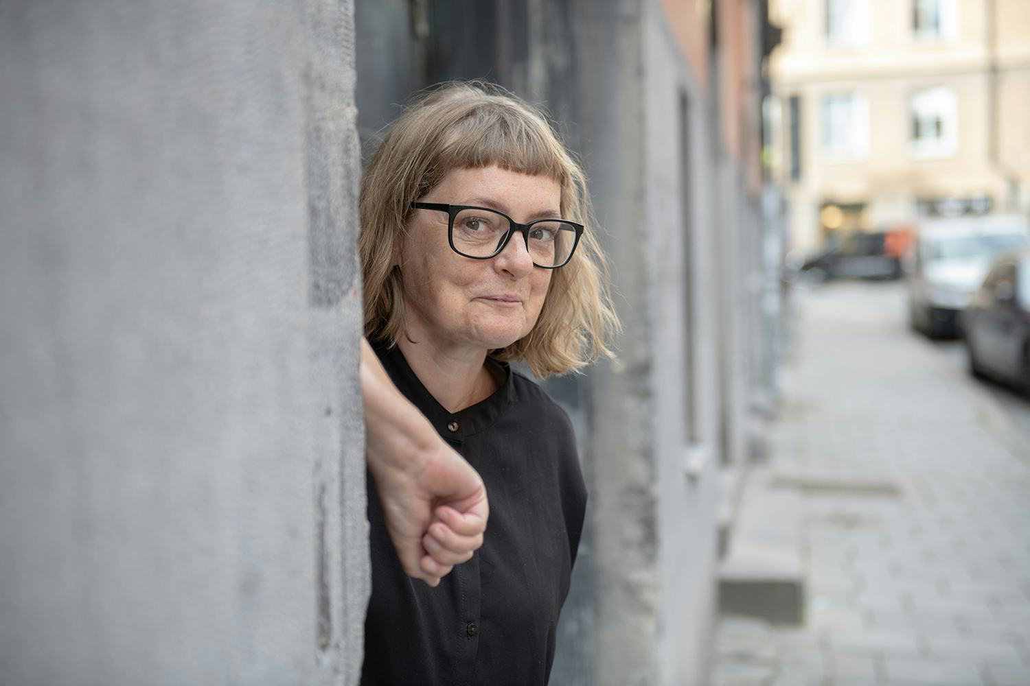 Amanda Creutzer tittar fram från grå vägg. Svata glasögon och skjorta. Ler finurligt.