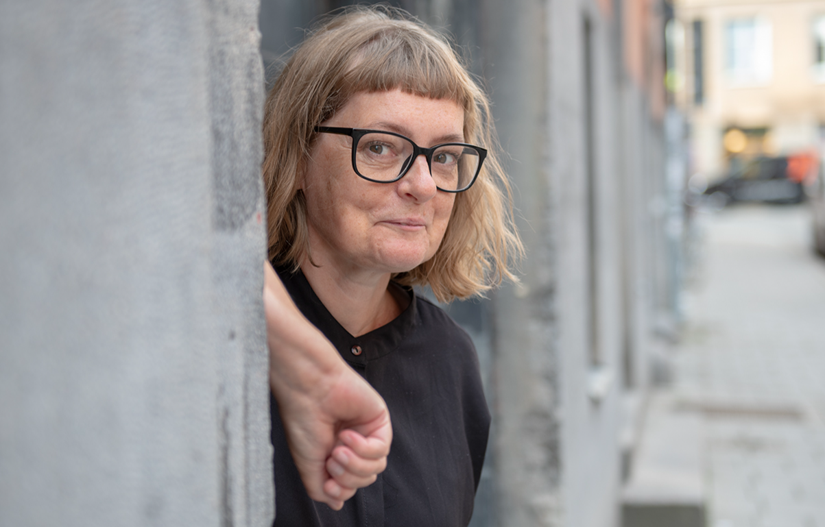 Amanda Creutzer tittar fram från grå vägg. Svata glasögon och skjorta. Ler finurligt.