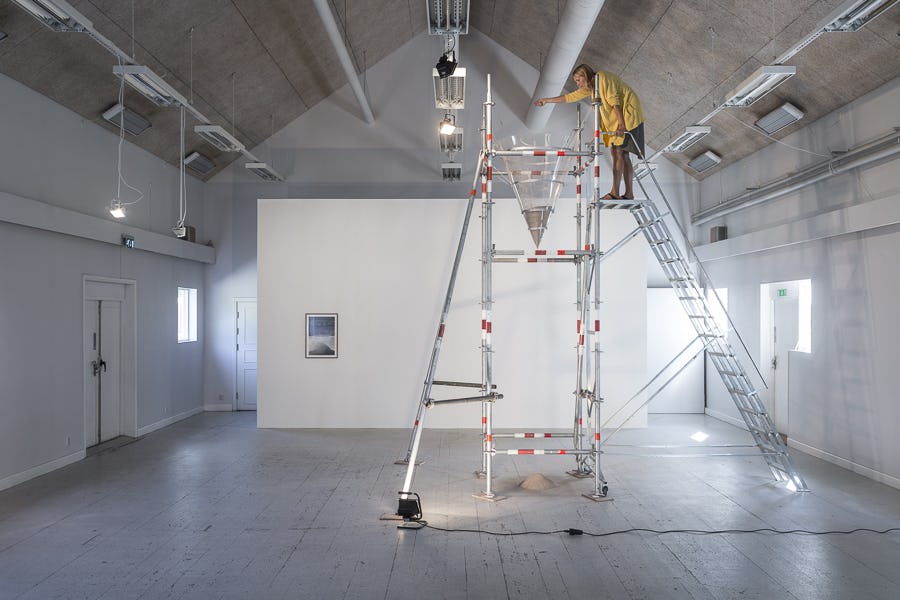 Kvinna med gul skjorta häller ner sand i en tratt, står på hög ställning med tillhörande trappa, i konsthallslokal.