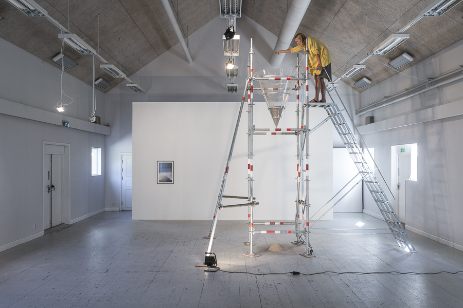 Kvinna med gul skjorta häller ner sand i en tratt, står på hög ställning med tillhörande trappa, i konsthallslokal.