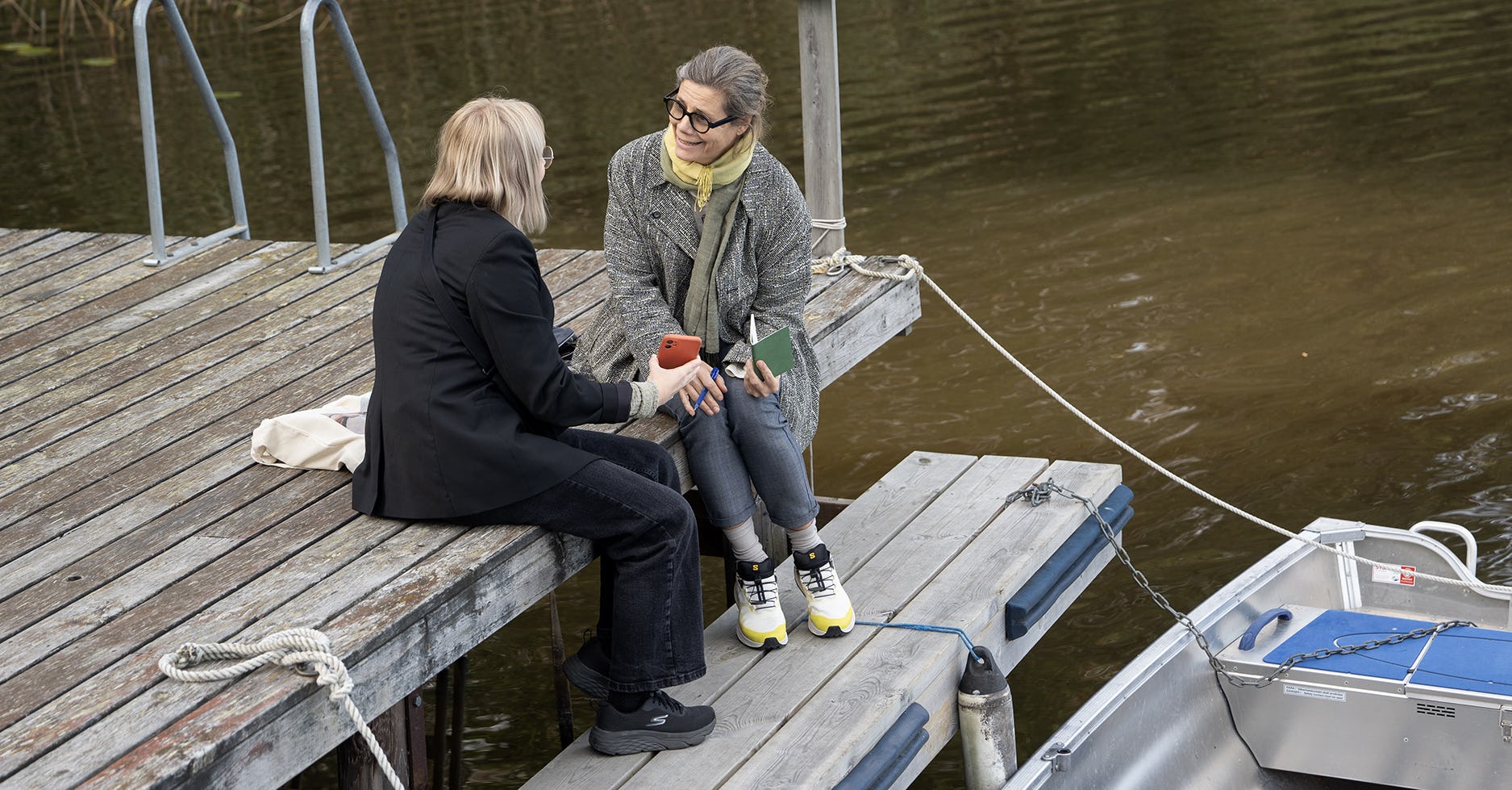 Två kvinnor pratar med varandra sittande på brygga.. Den ena visar sitt konstnärliga arbete på sin mobiltelefon.