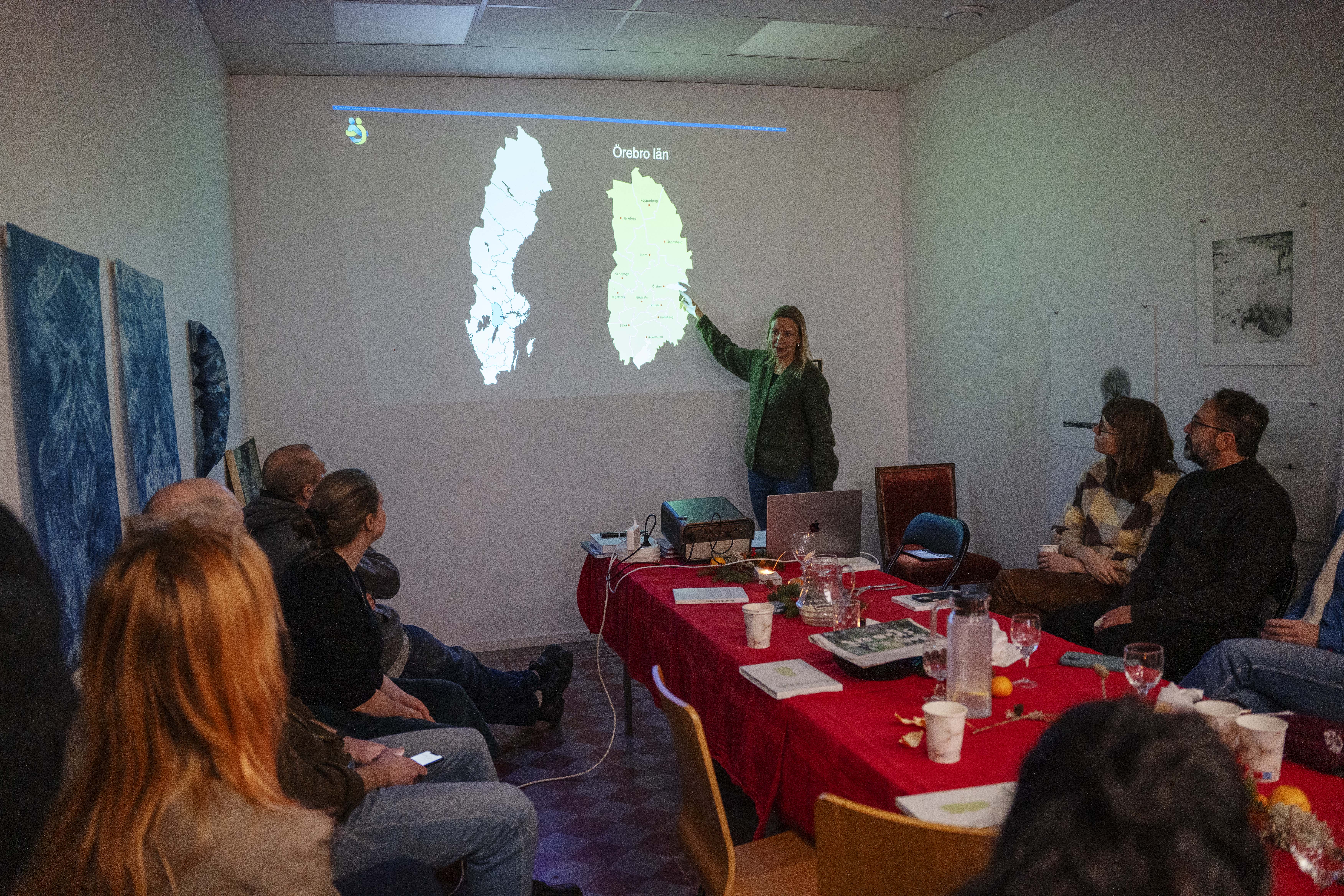 Kvinna föreläser och pekar på karta över Sverige och en region. Många deltagare sitter runt ett bord med julstämning, röd duk.