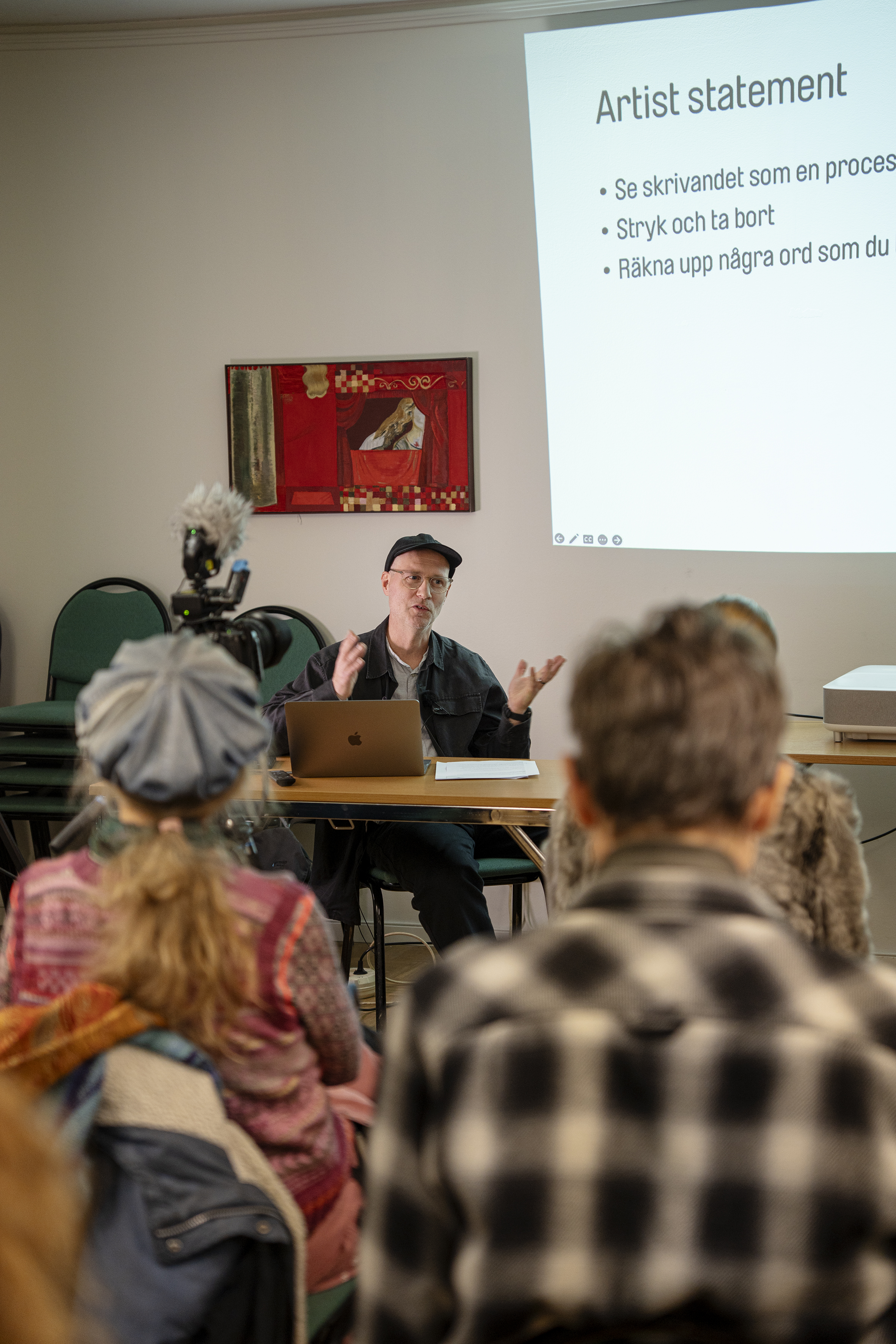 Föreläsaren Niklas Östholm gestikulerar i keps för publik, på väggen en röd tavla och projektion med orden Artist Statement.