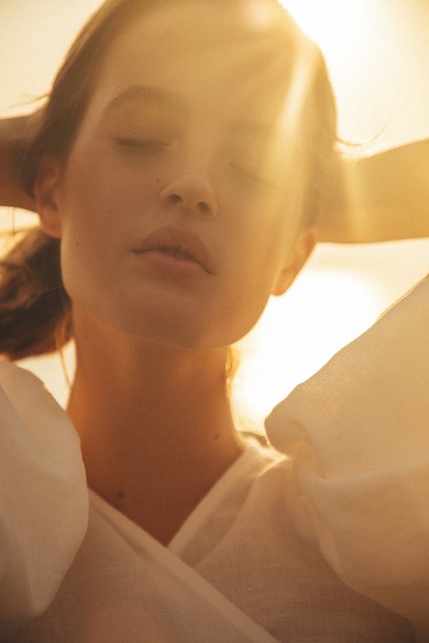 Woman with hands up at her pony tail, eyes closed while the sun glistens over her. Her firm and smooth skin show the potential results of peptide therapy X in Boston with Neem