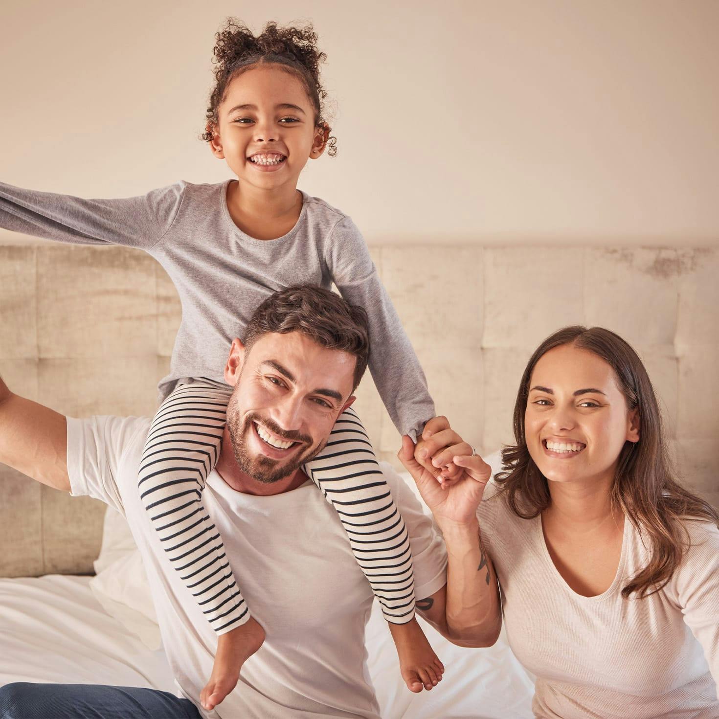 little girl sitting on her dad's shoulders next to her mom, sitting on a bed