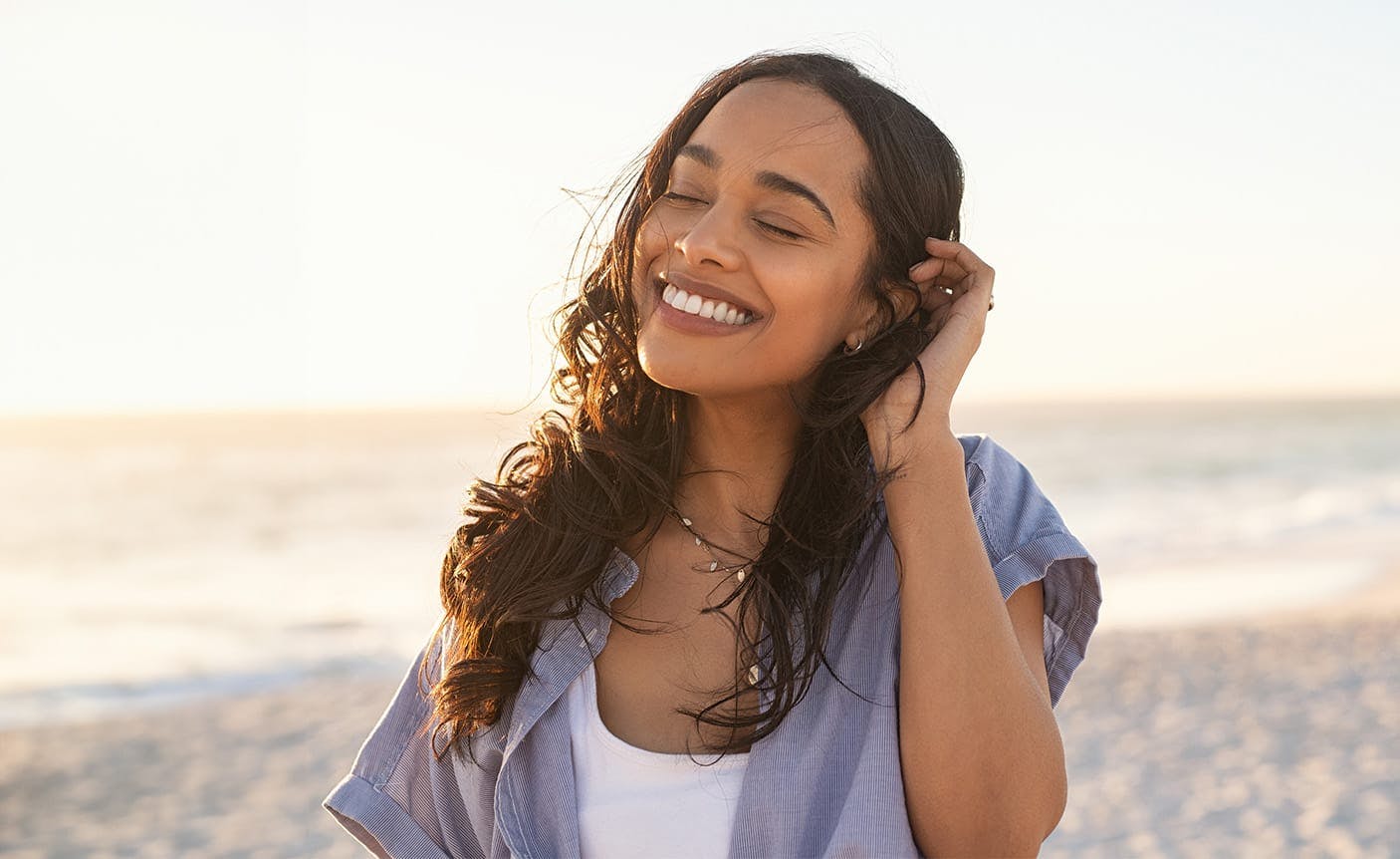 smiling woman brushing her hair behind her ear, standing on a beach