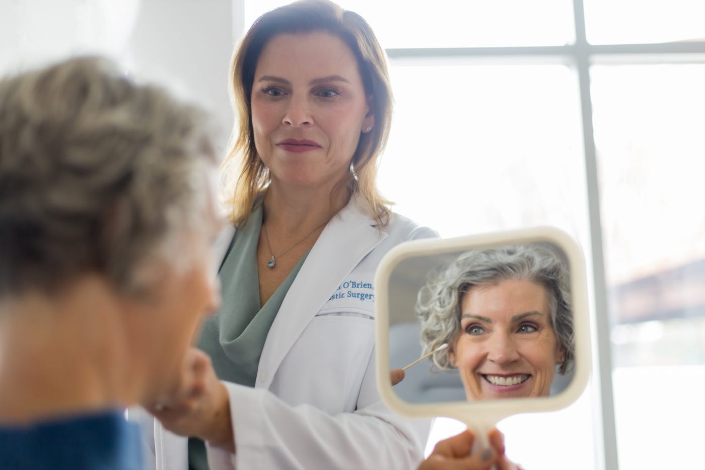 woman smiling into handheld mirror while doctor points to her brow bone