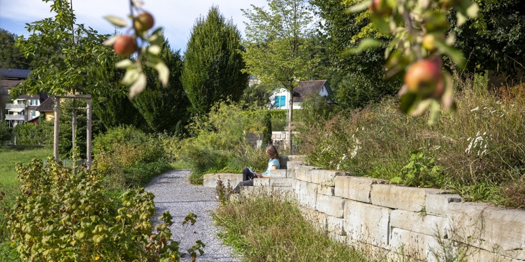 Frau ruht sich auf Steinbänkchen inmitten von Wildstauden und Obstbäumen aus.