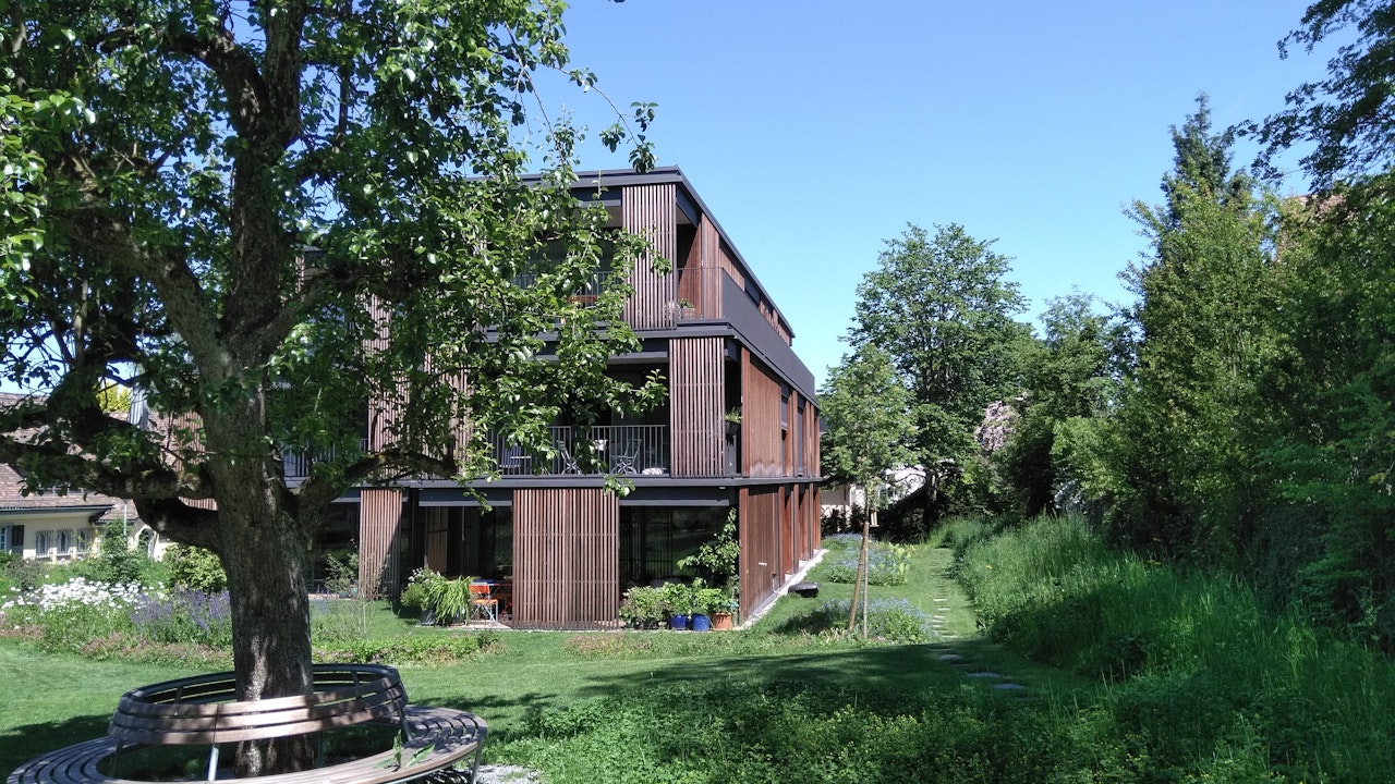 Schattenspendende Bäume, Sitzgelegenheiten und Blumenwiesen werten das Mehrfamilienhaus auf und schaffen ein naturnahes, lebenswertes Umfeld.