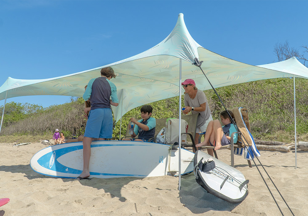 people under a shaded tent on the beach with a surf board