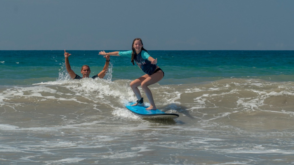 A teenager surfing in shallow water and another person cheering