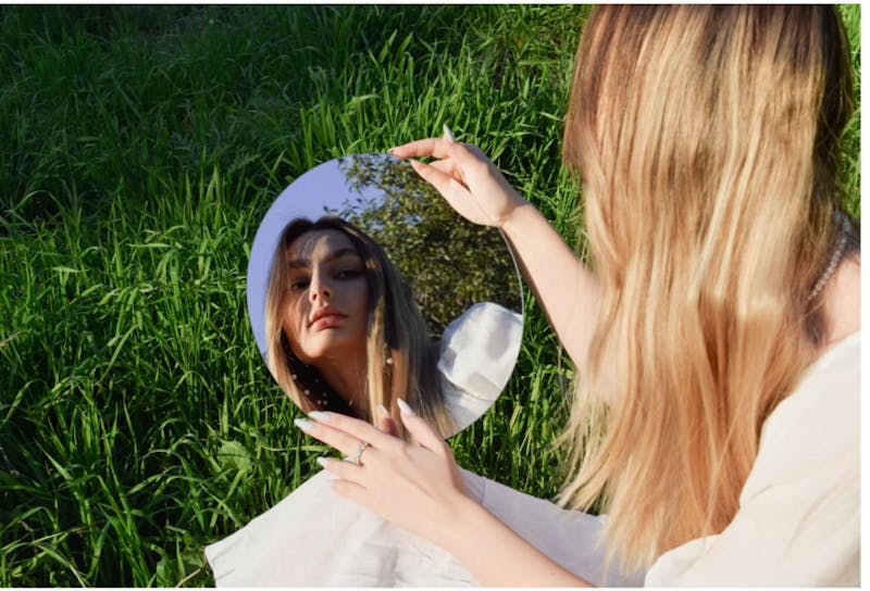 Woman sitting in spring grass looking at her reflection in a mirror