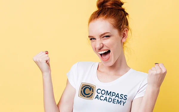 En begejstret rødhåret ung kvinde med en hvid t-shirt på med et Compass Academy logo