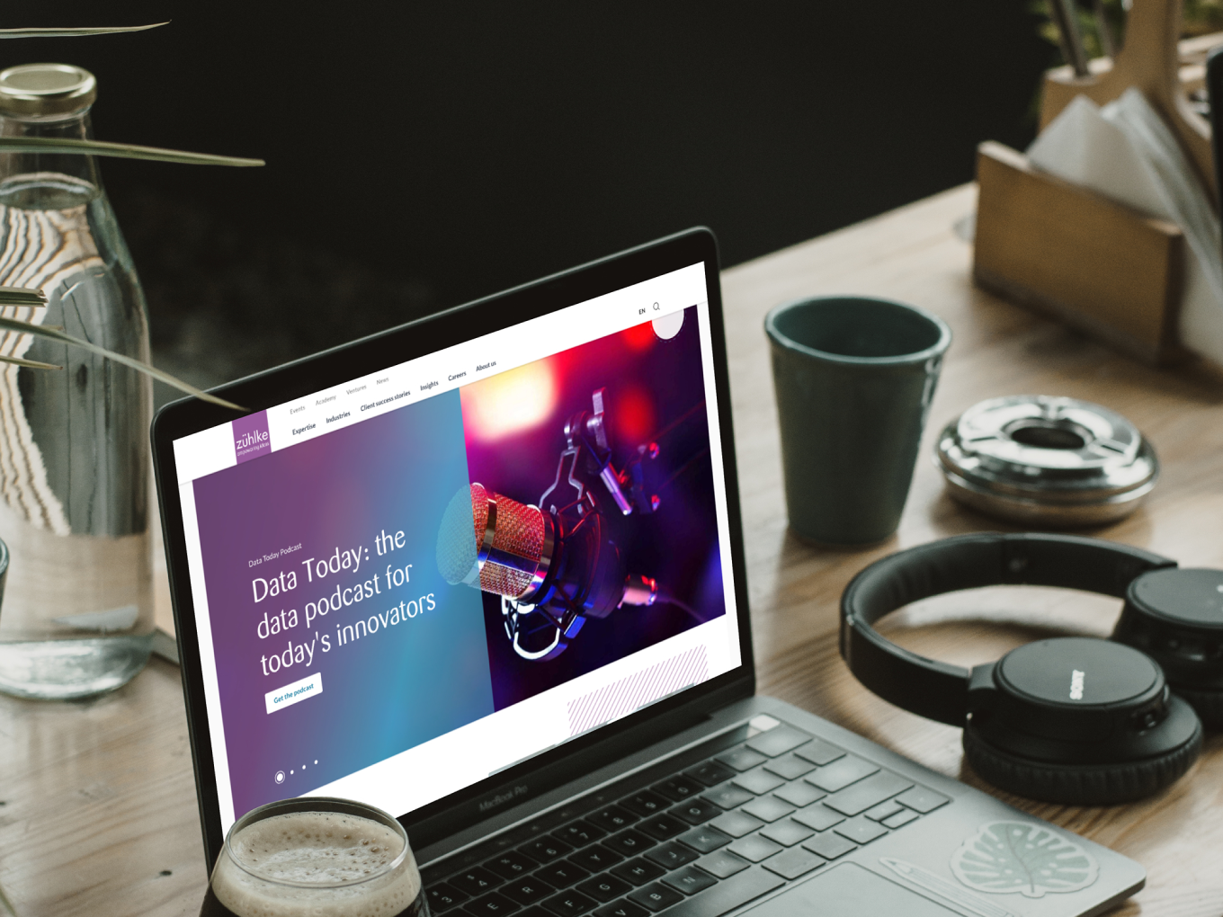 A laptop on a table displays a website with a podcast titled "Data Today: the data podcast for today's innovators." Nearby are headphones, a coffee cup, and a water bottle.