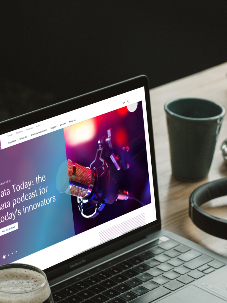 A laptop on a table displays a website with a podcast titled "Data Today: the data podcast for today's innovators." Nearby are headphones, a coffee cup, and a water bottle.