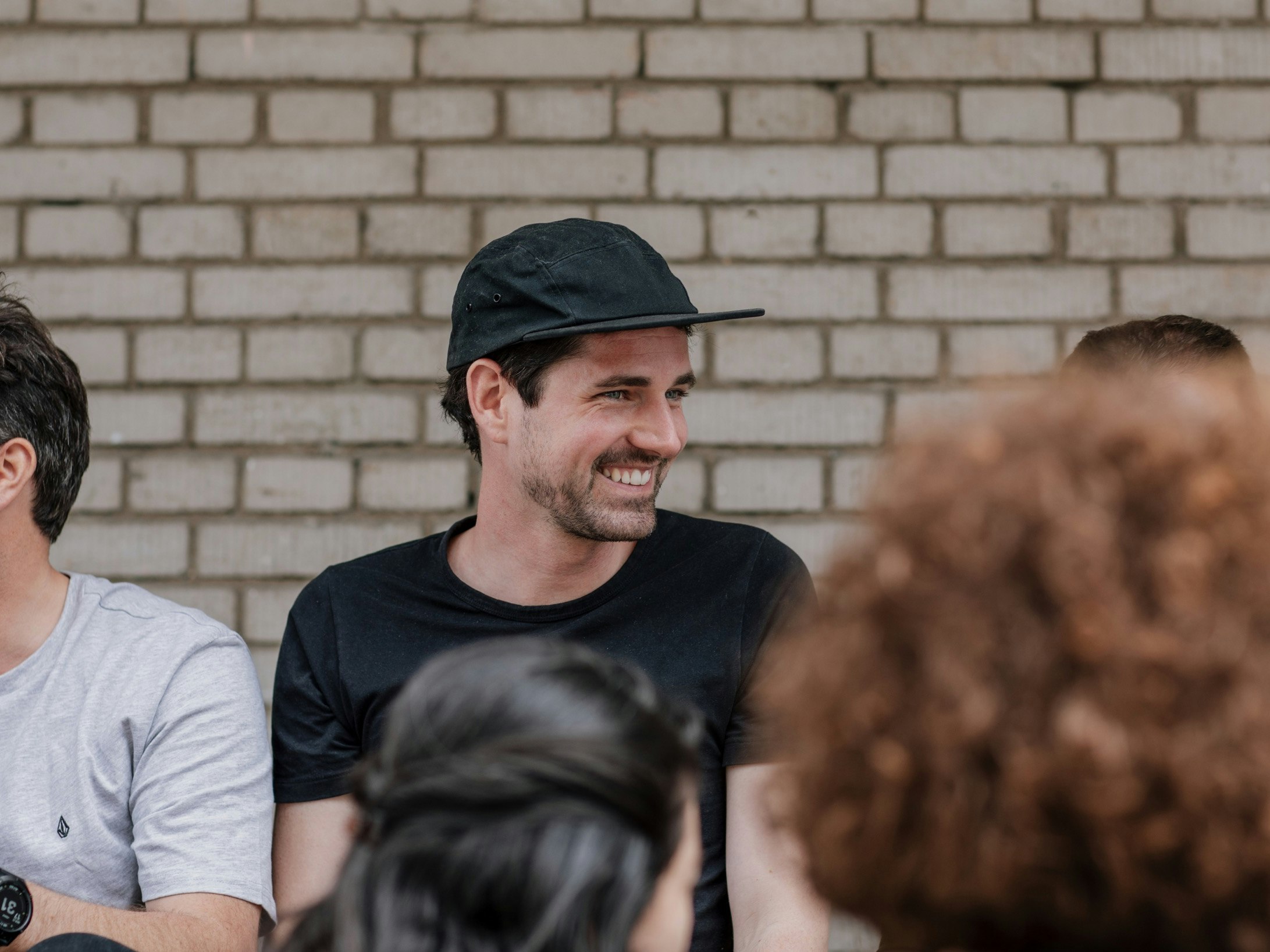 A man in a black cap and t-shirt smiles while sitting against a brick wall, surrounded by others.