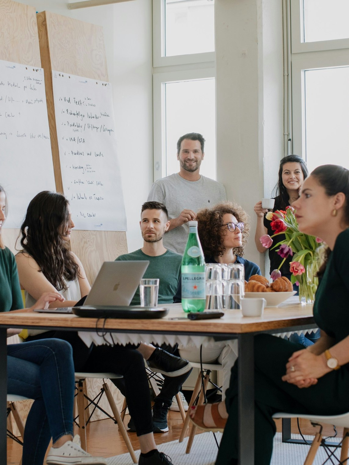 A group of people sitting and standing around a table in a bright room with large windows and flip charts.