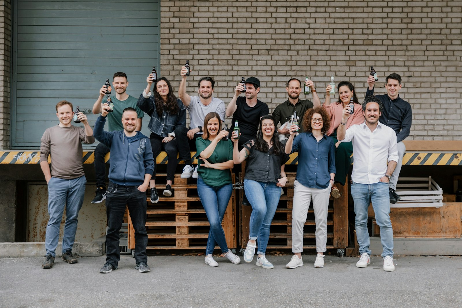 A group of 15 people, some standing and some sitting on wooden pallets, holding drinks and posing in front of a brick wall.