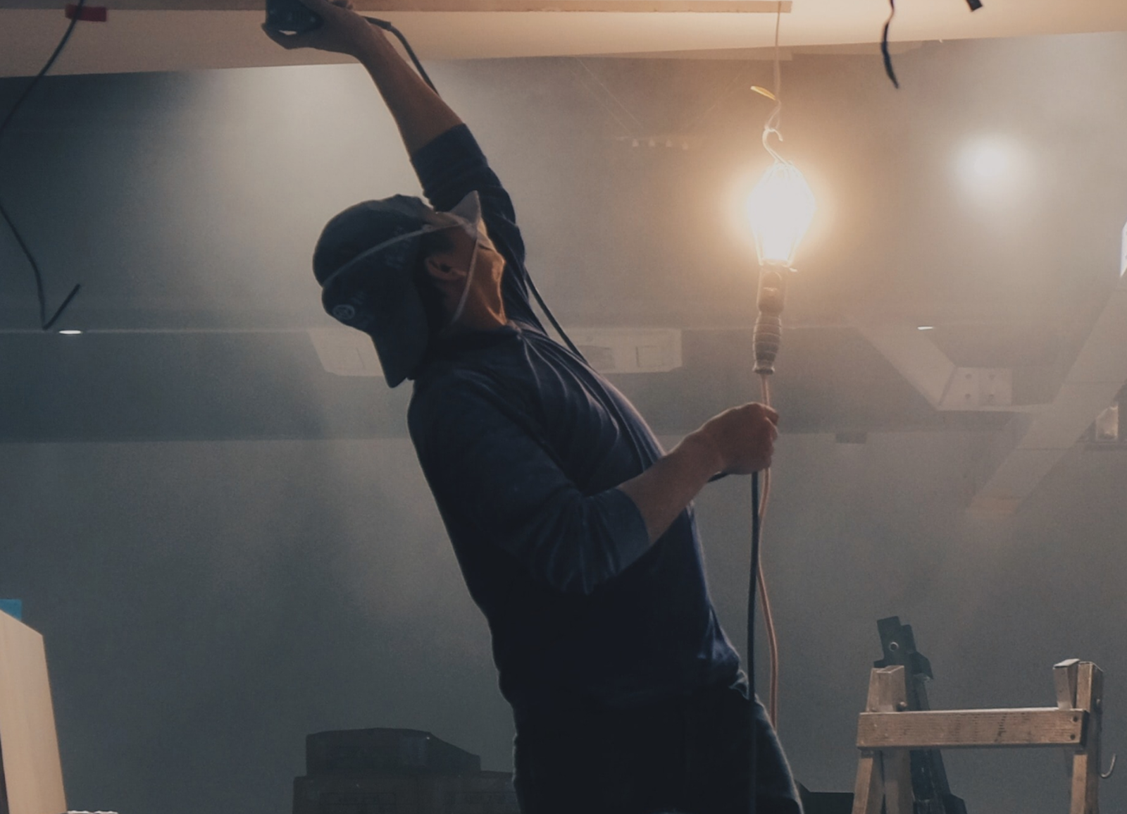 Person wearing protective gear working on a ceiling, next to a lit bulb, using a tool. A ladder is visible nearby.