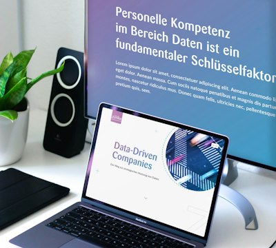 Close-up of a laptop and monitor displaying presentations about data-driven companies. A potted plant and speakers are in the background.