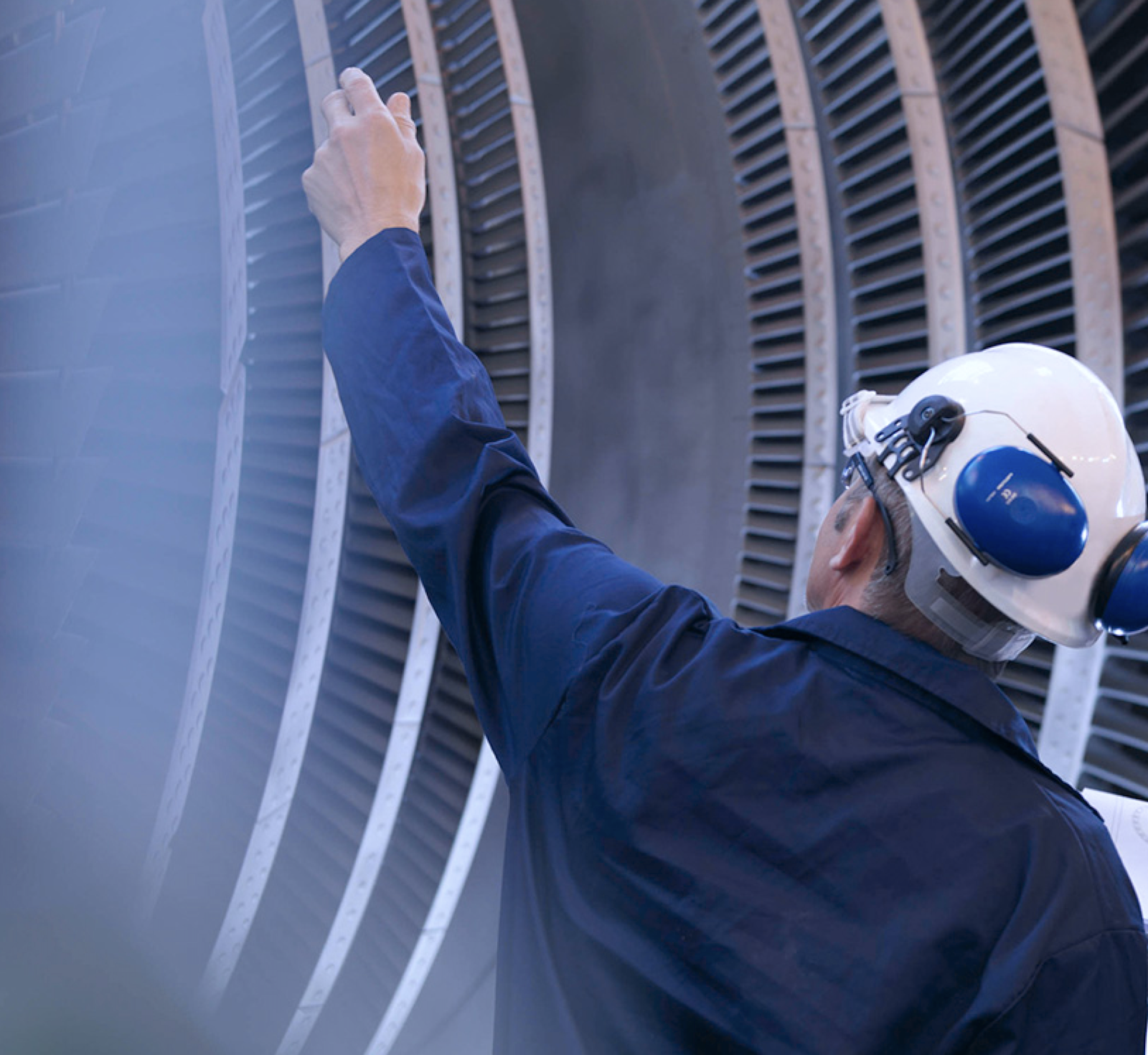 A worker in a hard hat and safety gear inspects a large industrial turbine, pointing at its blades.