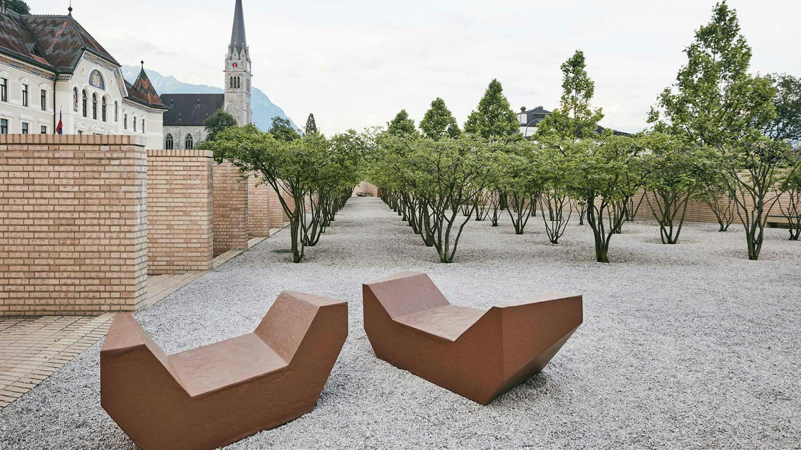 Two modern, angular benches on a gravel area surrounded by rows of trees, with a church and brick buildings in the background.