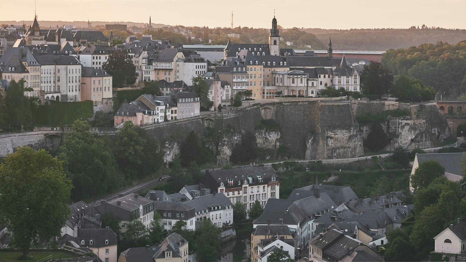 Aerial view of a historic cityscape with a mix of old buildings, a church, and greenery, set on a hill with fortified walls under a cloudy sky.