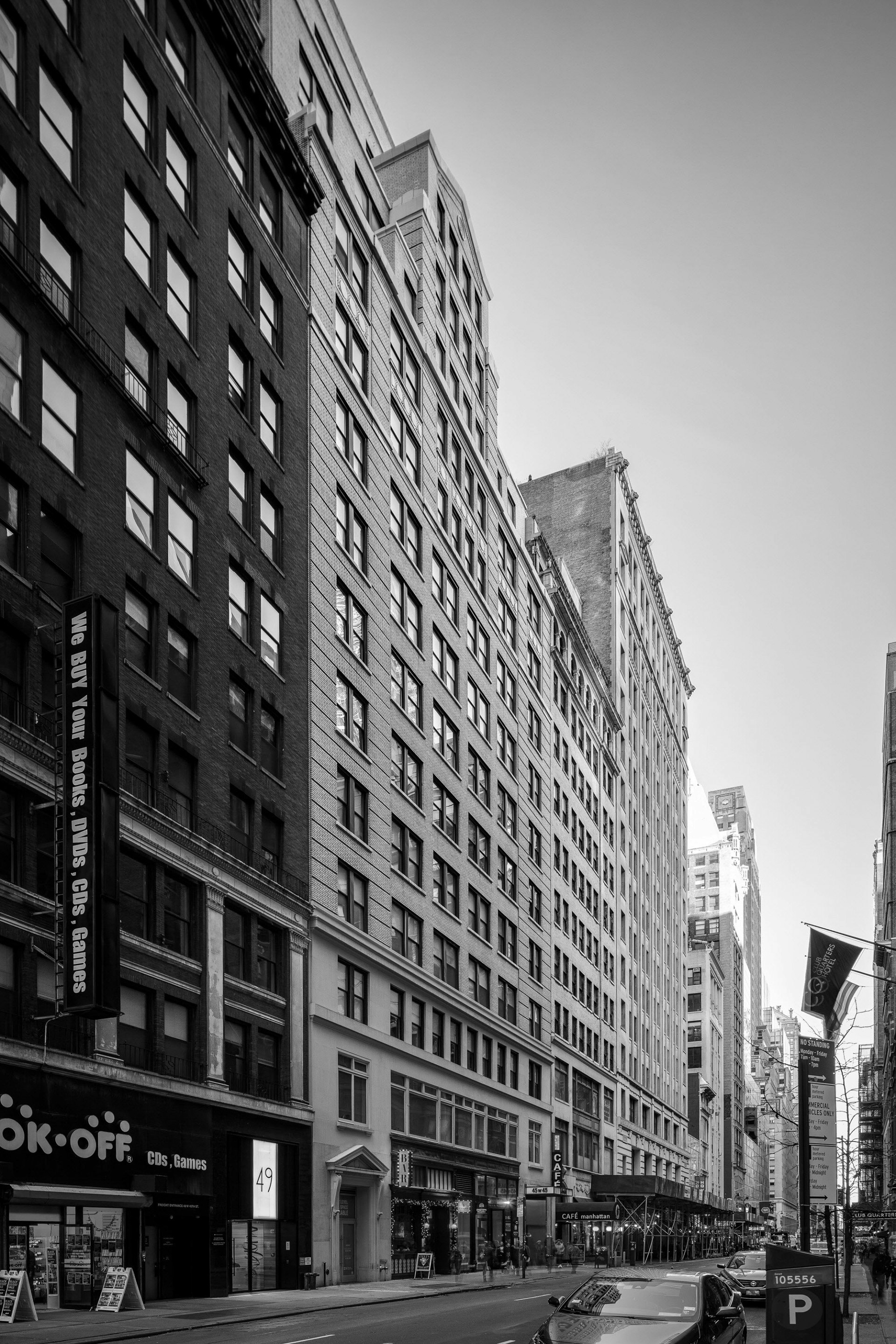 Black and white photo of a city street with tall buildings lining both sides. Signs for shops, cars parked along the road, and pedestrians are visible.