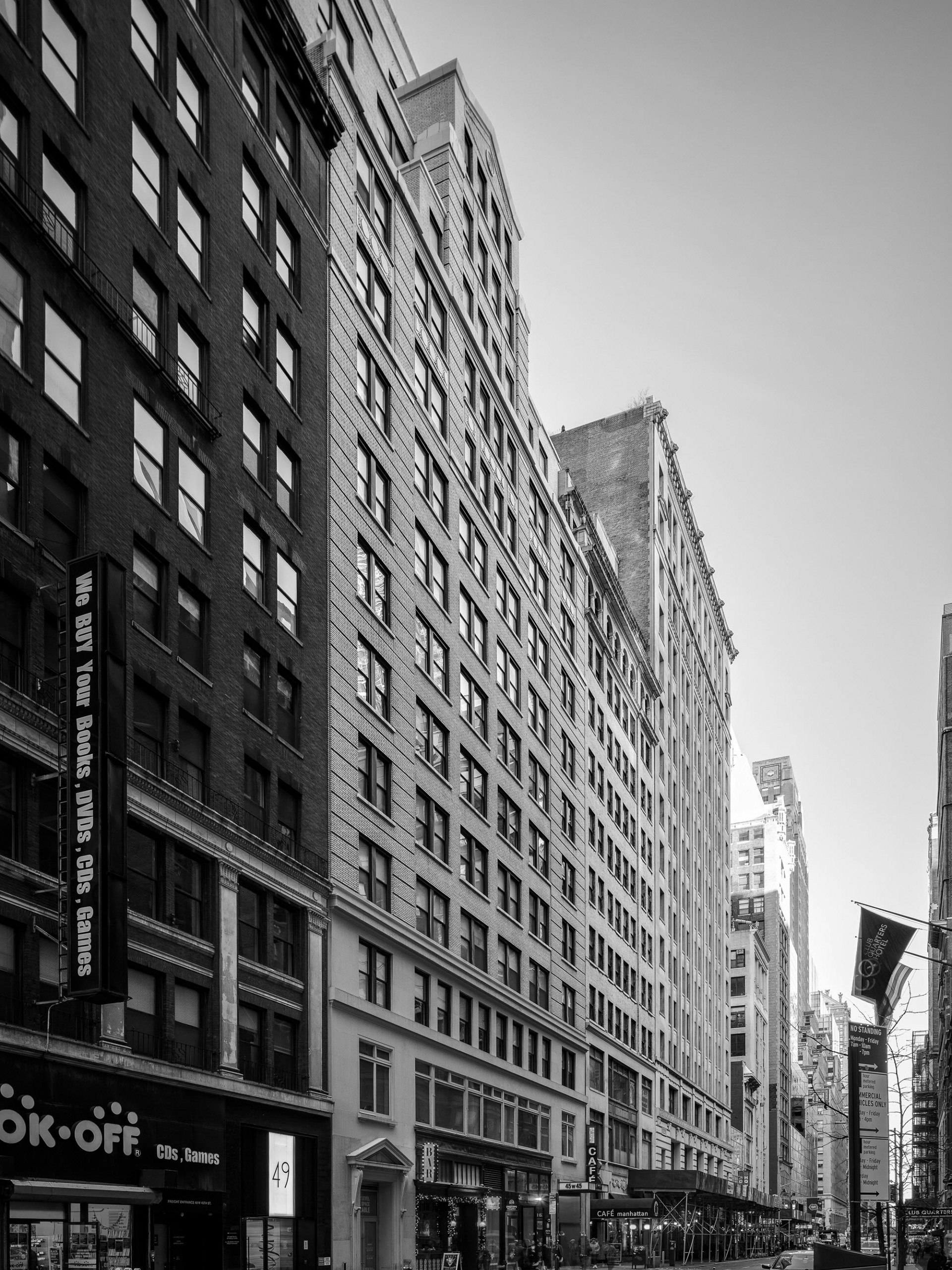 Black and white photo of a city street with tall buildings lining both sides. Signs for shops, cars parked along the road, and pedestrians are visible.