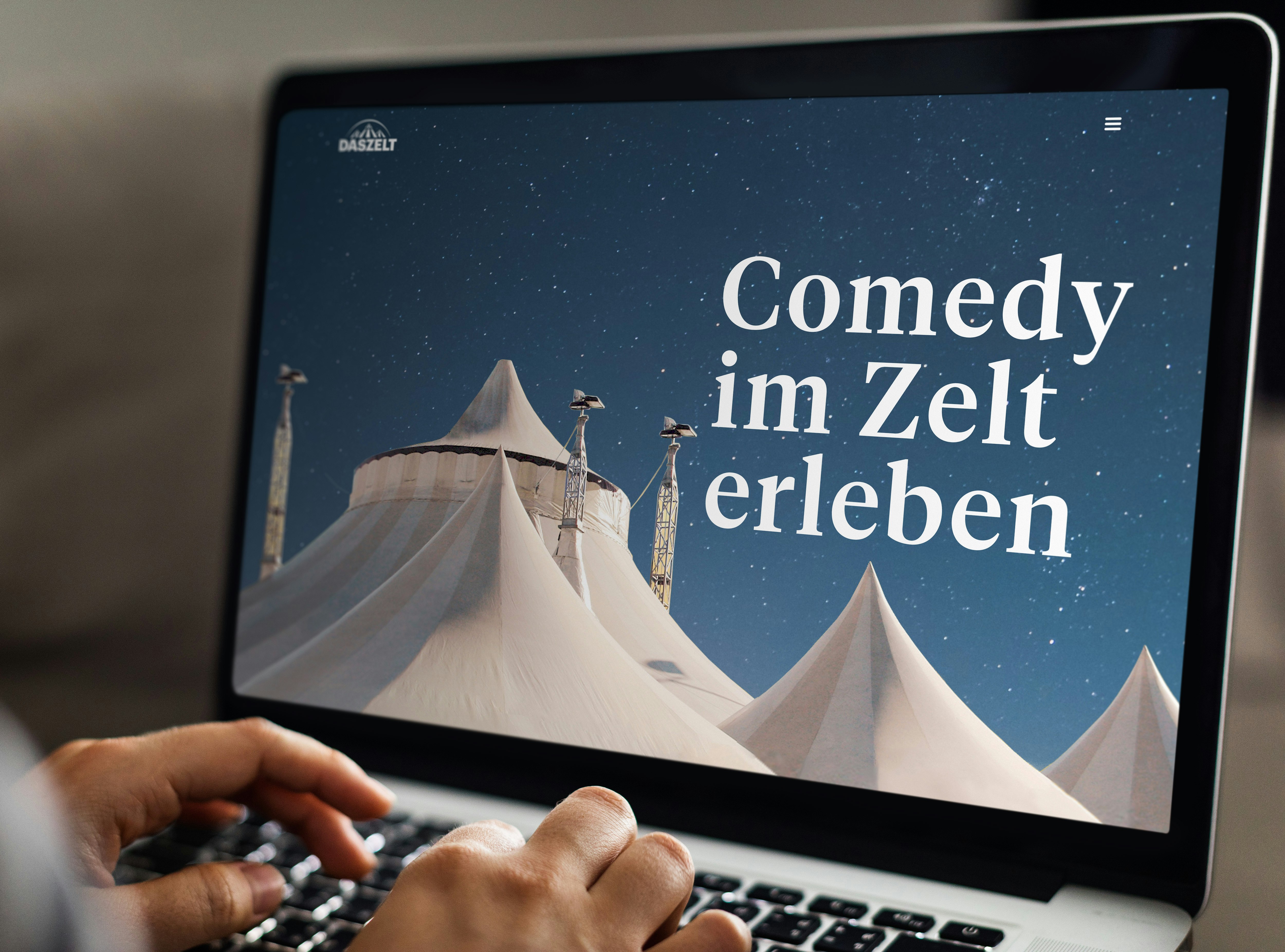 A person types on a laptop displaying a webpage with white tents under a starry sky. The text reads "Comedy im Zelt erleben.