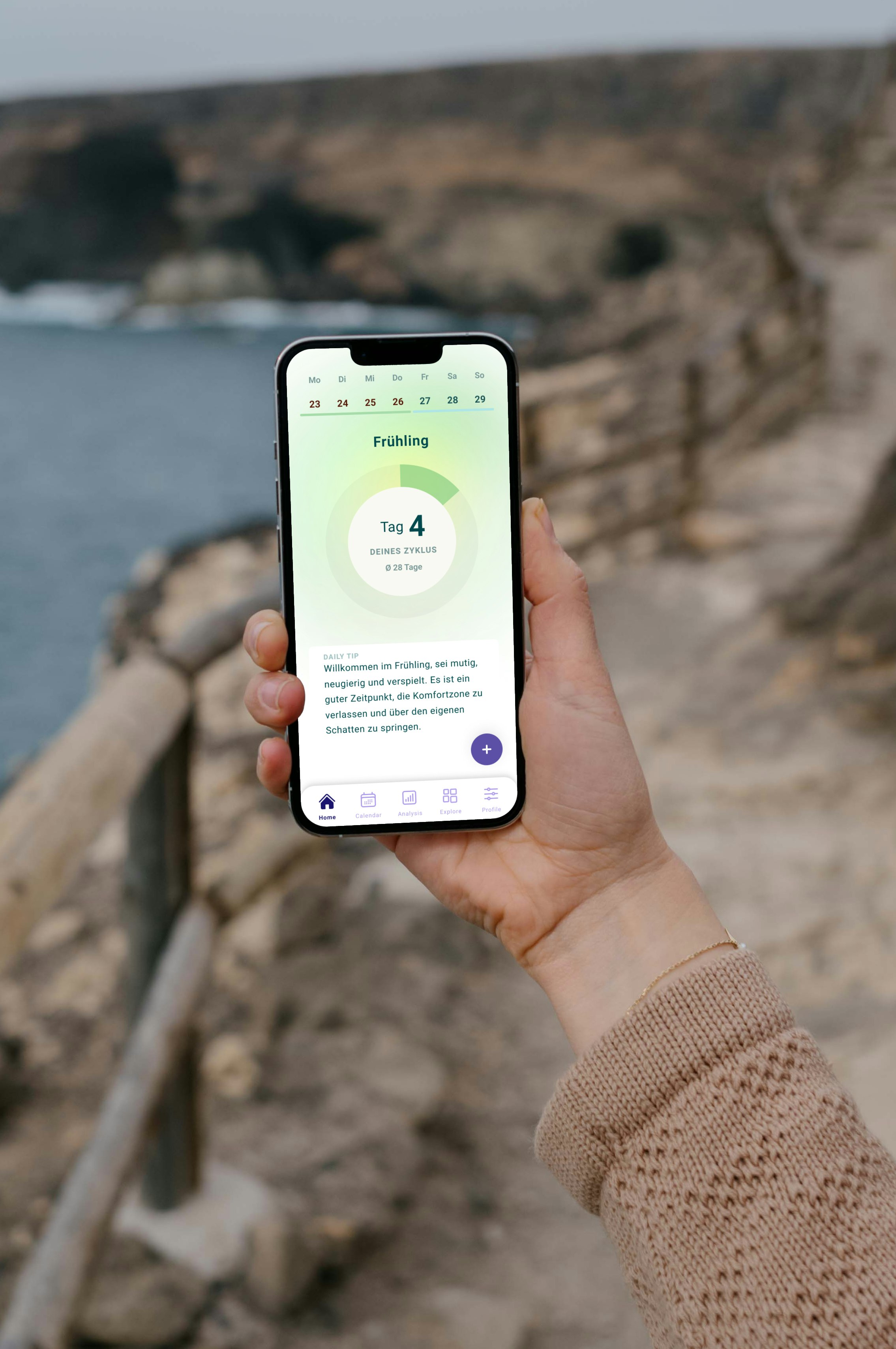 Person holding a smartphone displaying a weather app with the day marked as "Frühling Tag 4," standing on a pathway next to a rocky coastline.