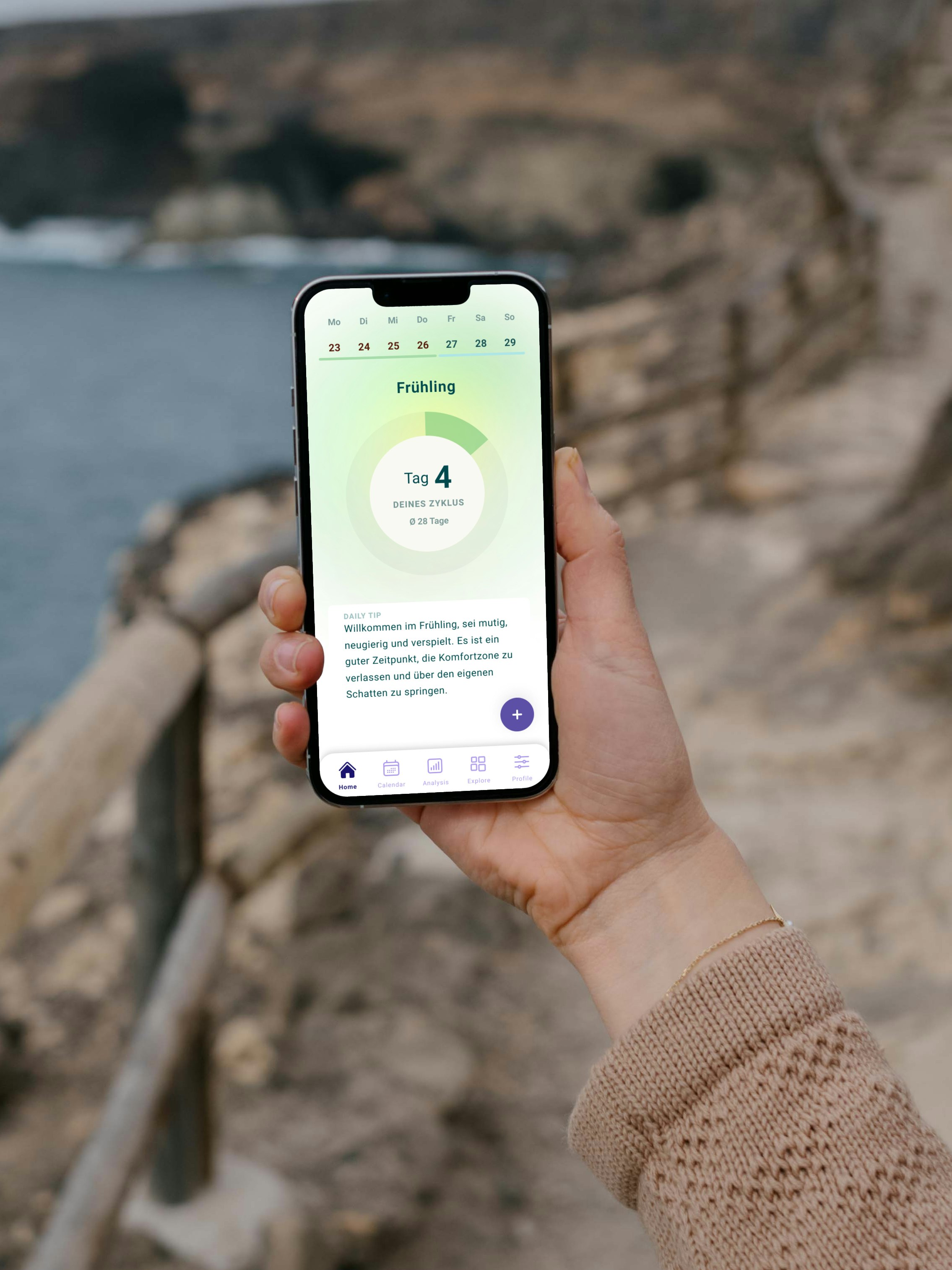 Person holding a smartphone displaying a weather app with the day marked as "Frühling Tag 4," standing on a pathway next to a rocky coastline.