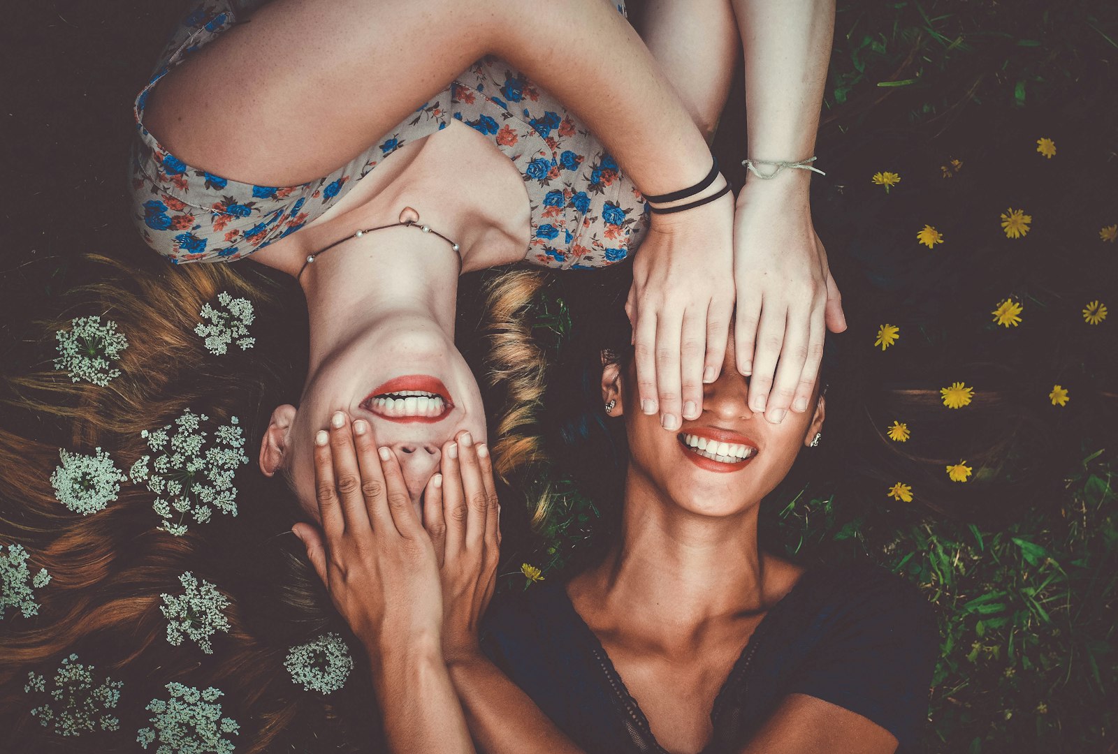 Two women lie on grass, smiling with eyes covered by each other's hands, surrounded by flowers.