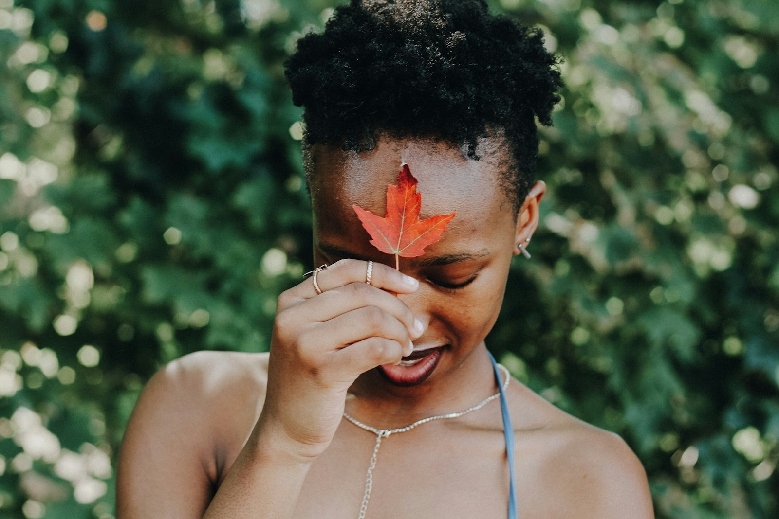 Person holds a red maple leaf on their forehead, standing against a blurred green foliage background.