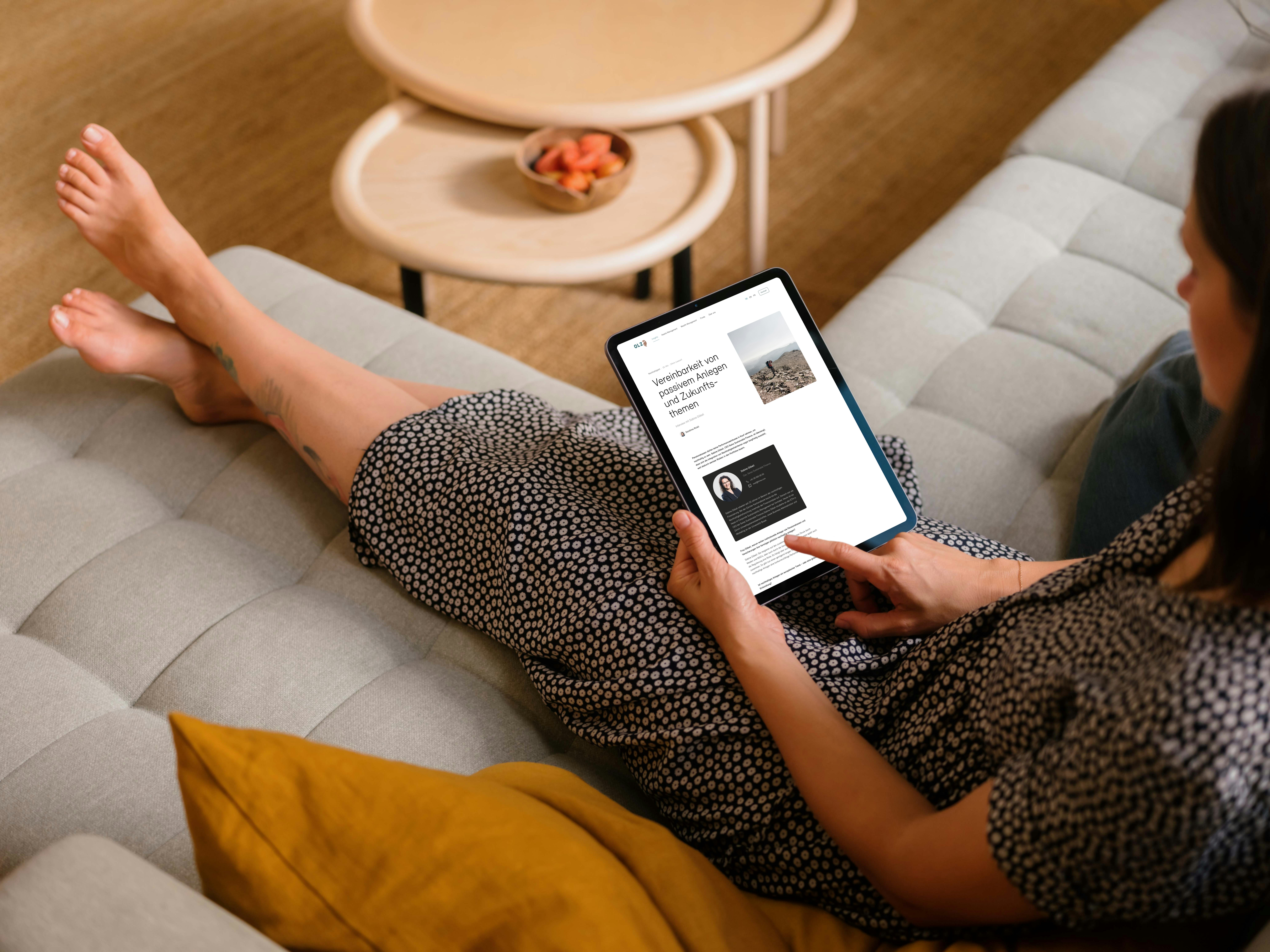 A person in a patterned dress sits on a sofa, using a tablet. Two round tables with items are in the background.