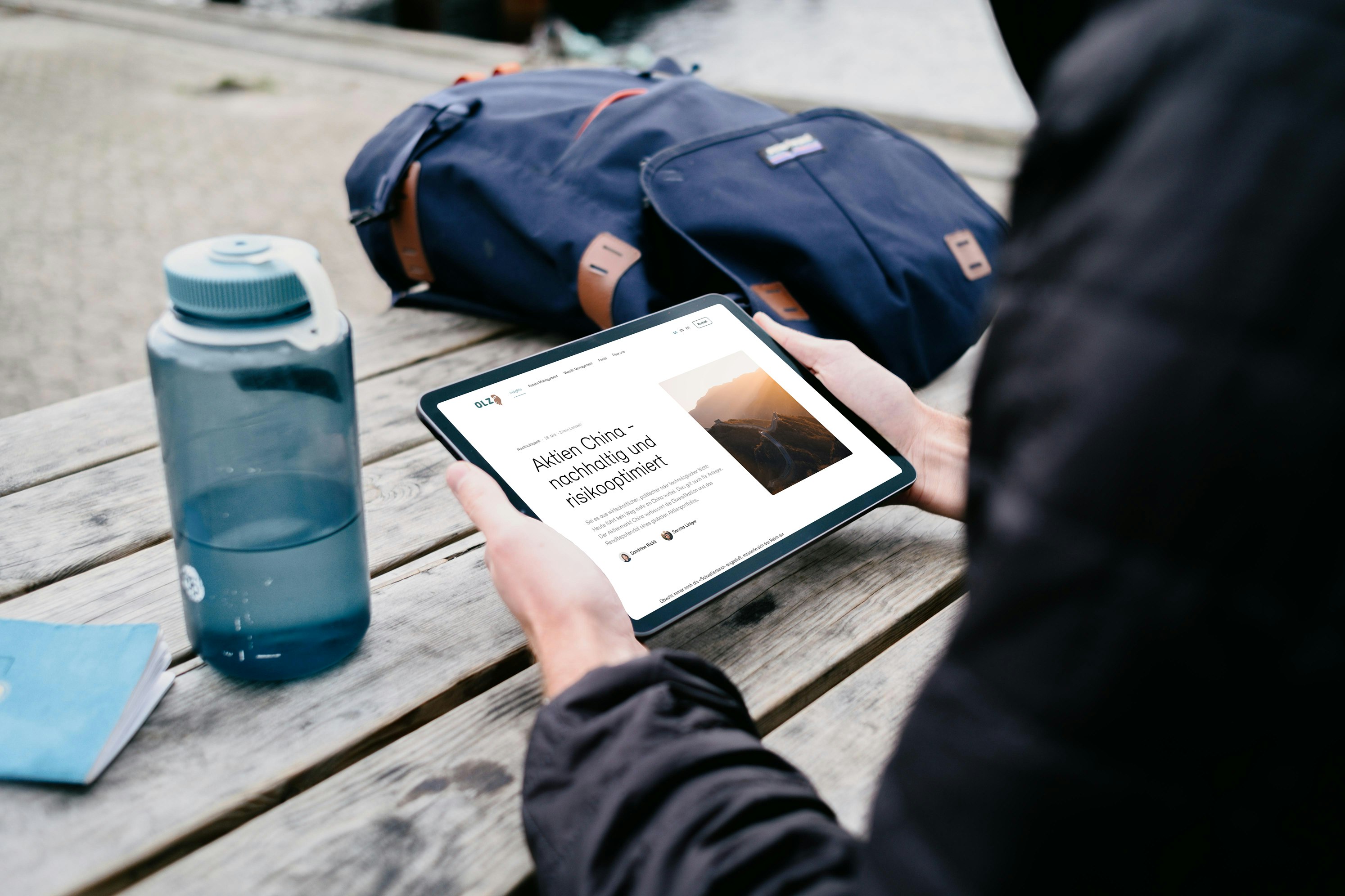 Person seated at a picnic table using a tablet, with a bottle and a backpack nearby.