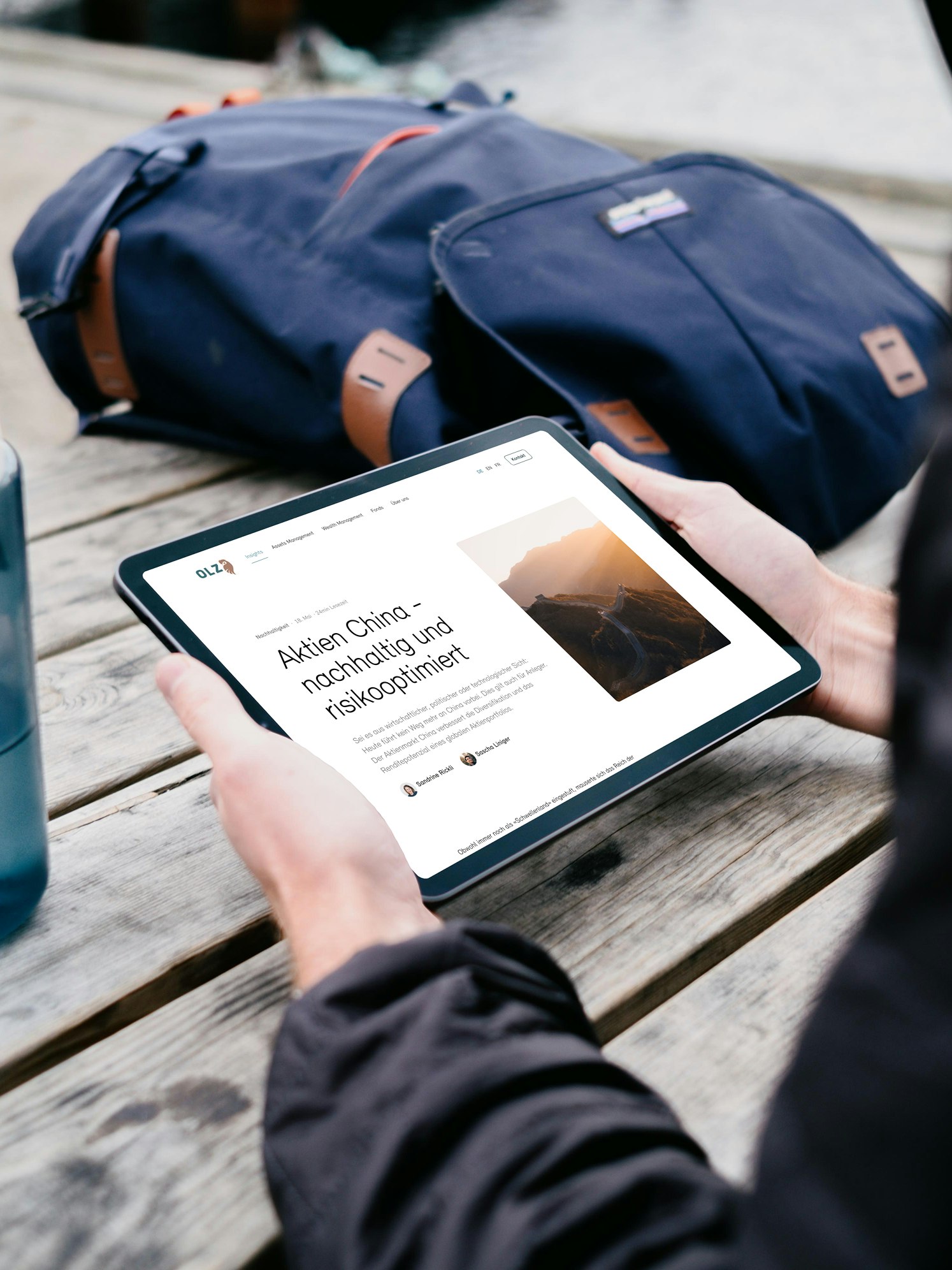 Person seated at a picnic table using a tablet, with a bottle and a backpack nearby.
