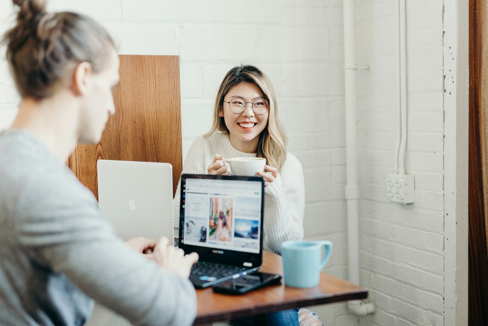 Two people sit at a table with laptops. The person on the left is focused on the computer, while the person on the right smiles and holds a cup.