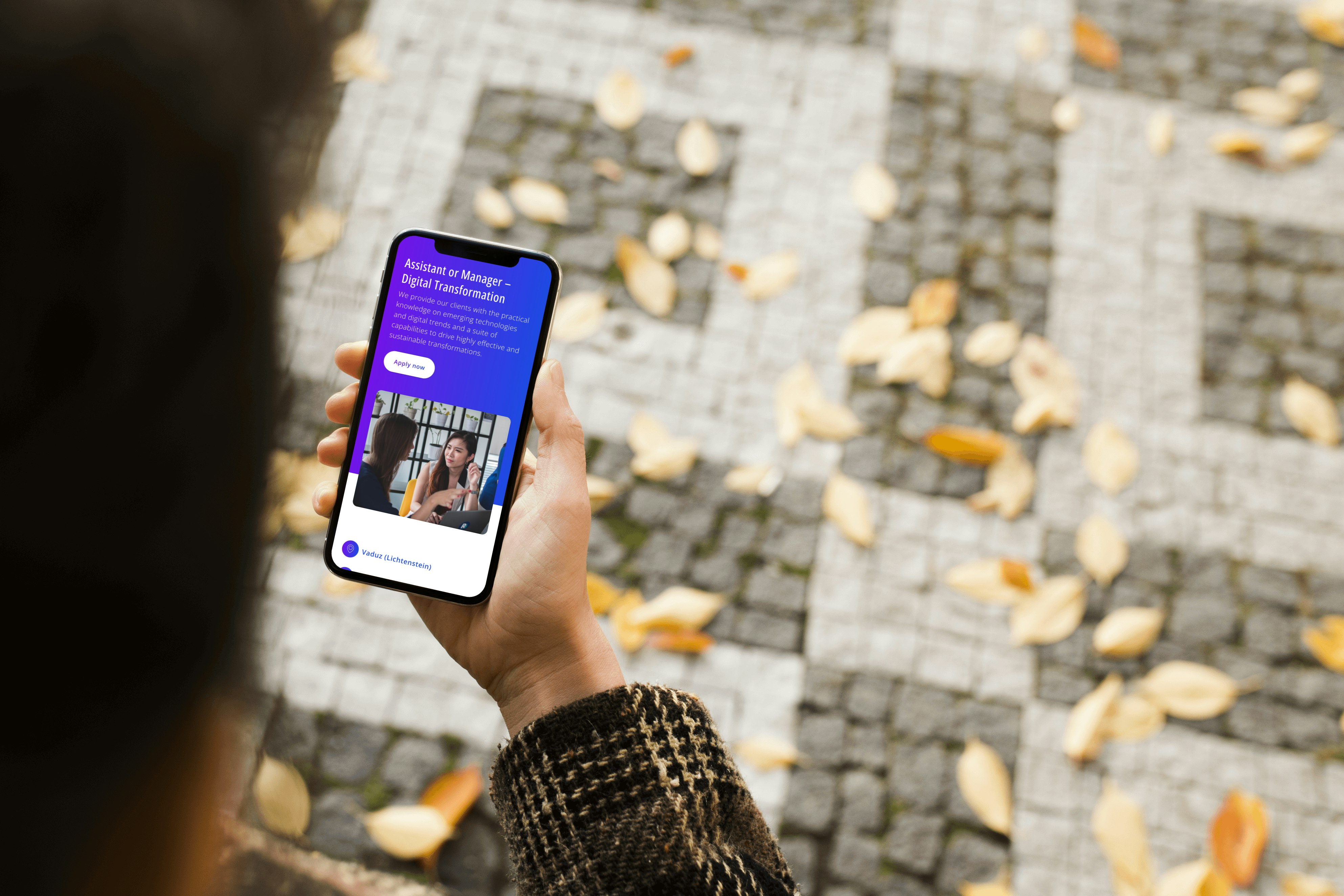 Person holding a smartphone displaying a digital transformation app interface, with autumn leaves scattered on a cobblestone path in the background.