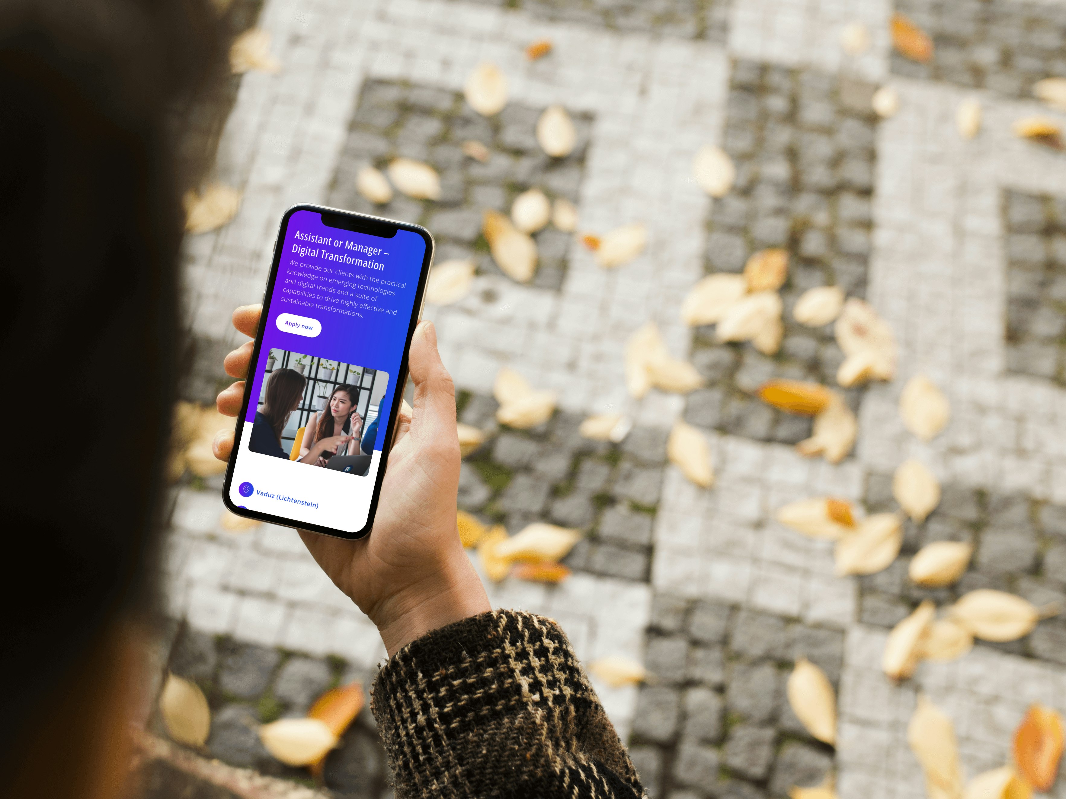 Person holding a smartphone displaying a digital transformation app interface, with autumn leaves scattered on a cobblestone path in the background.