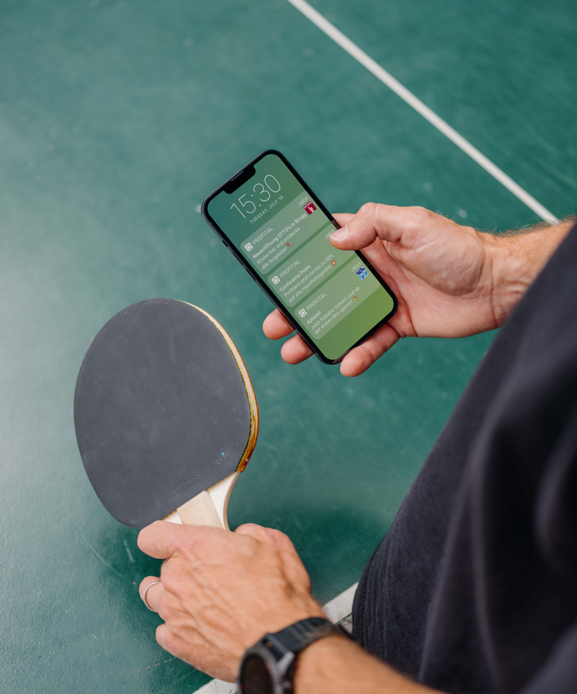 Person holding a smartphone displaying notifications in one hand and a table tennis paddle in the other, standing on a green court surface.