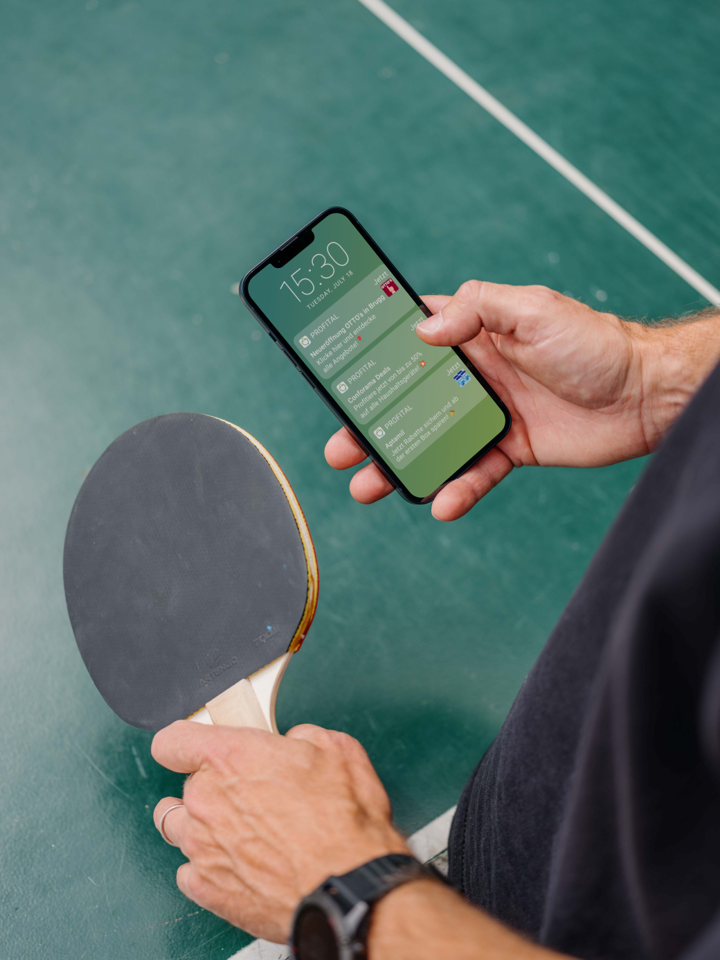 Person holding a smartphone displaying notifications in one hand and a table tennis paddle in the other, standing on a green court surface.