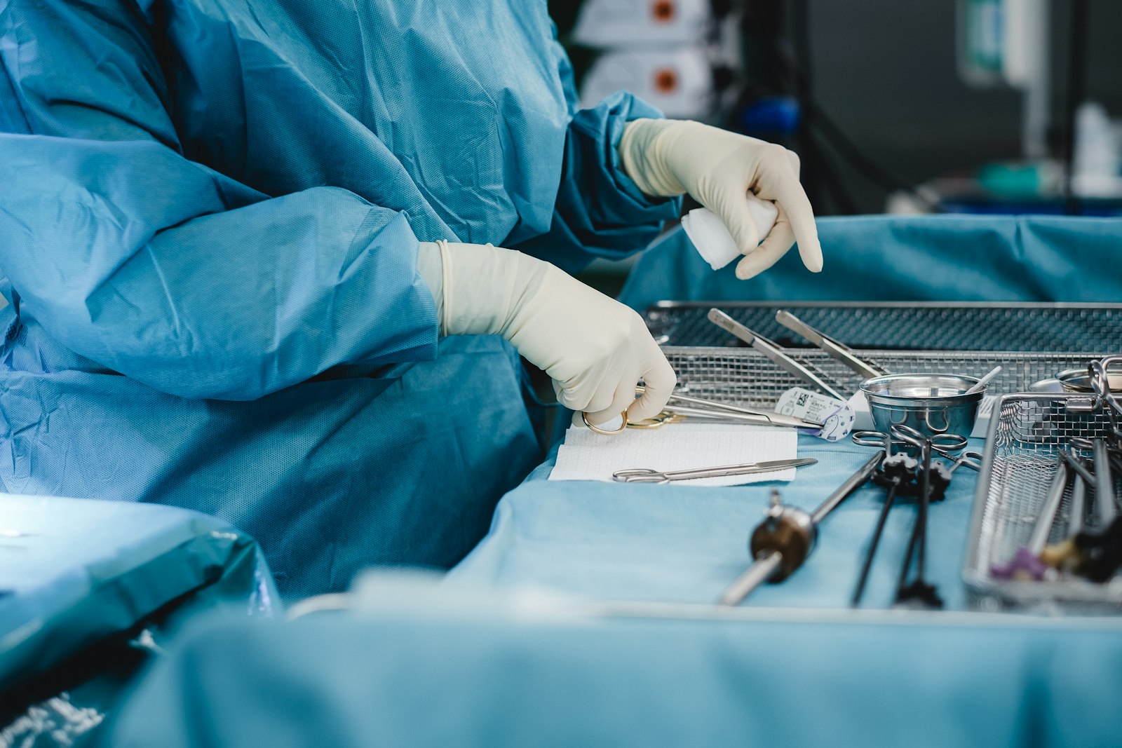 A surgeon in blue scrubs and gloves prepares surgical instruments on a table in an operating room.