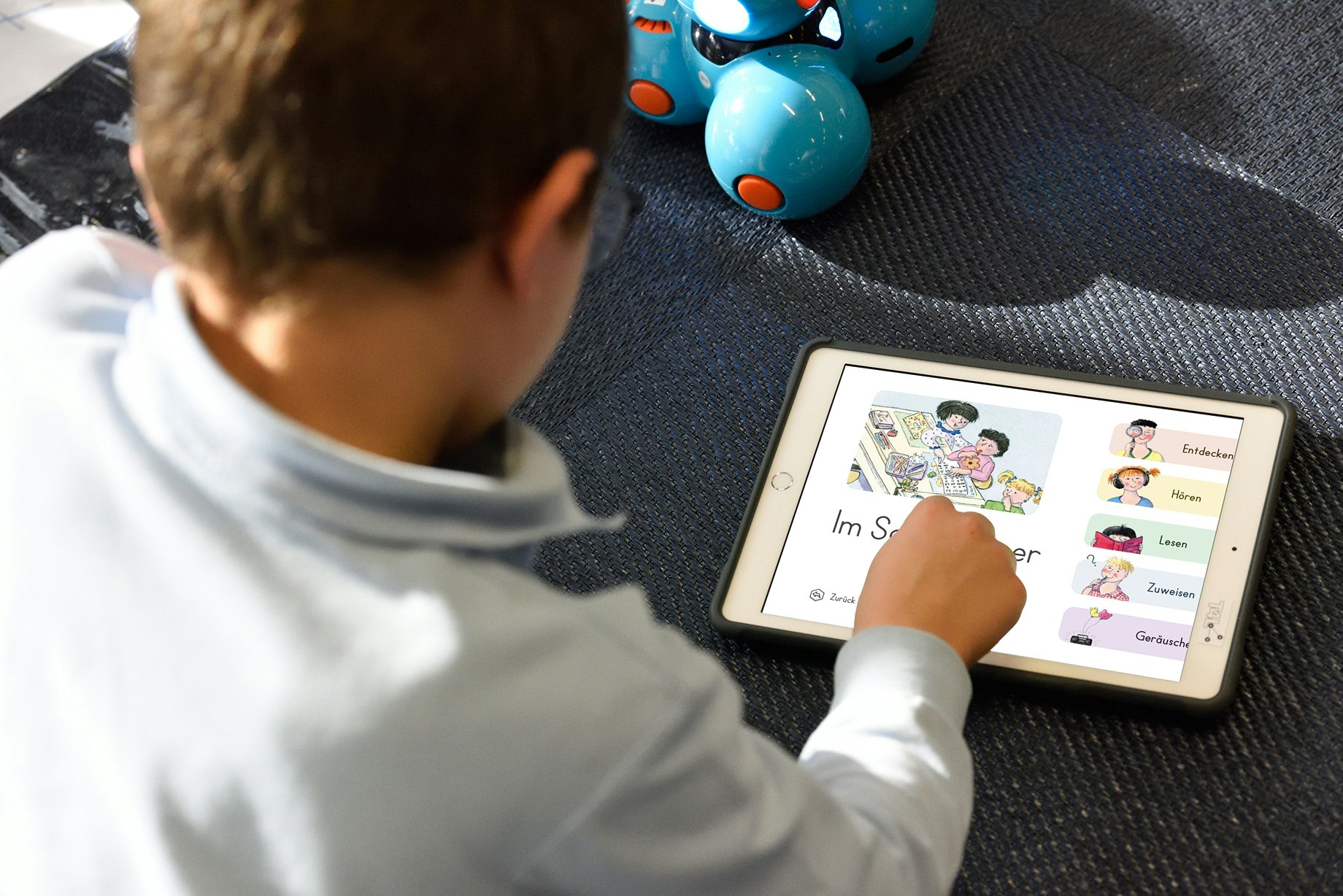 A child uses a tablet with a German language app. Blue robotic toys are visible in the background.