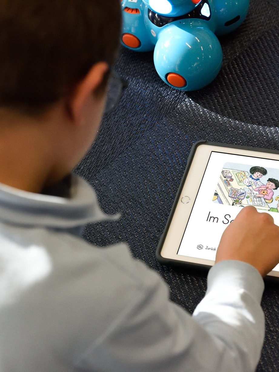 A child uses a tablet with a German language app. Blue robotic toys are visible in the background.