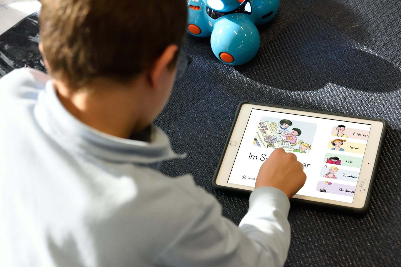 A child uses a tablet with a German language app. Blue robotic toys are visible in the background.