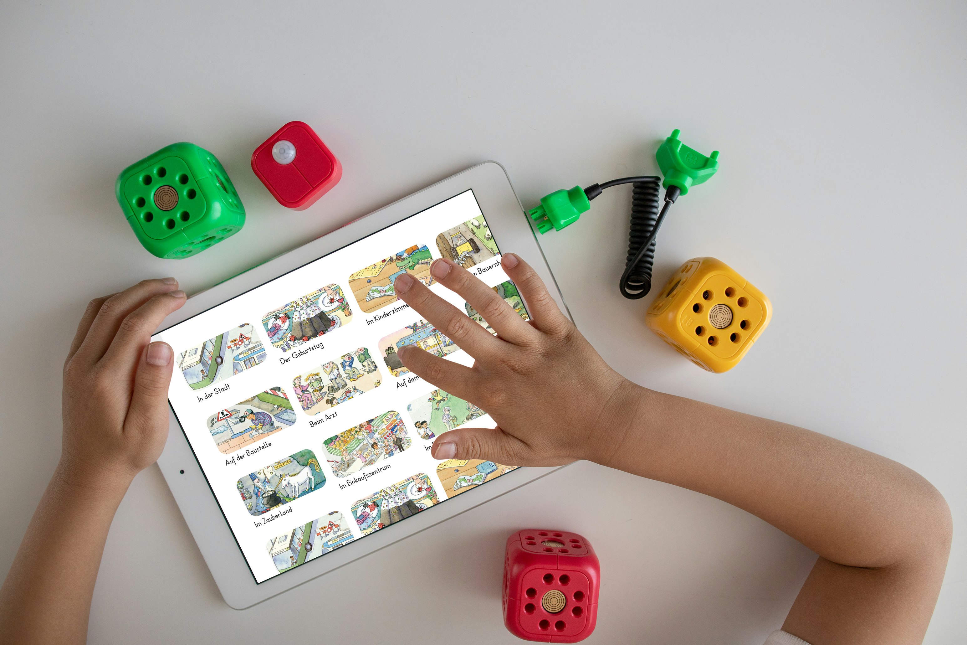 A child's hand interacts with a tablet displaying illustrated stories, surrounded by colorful plastic blocks and connectors on a white surface.