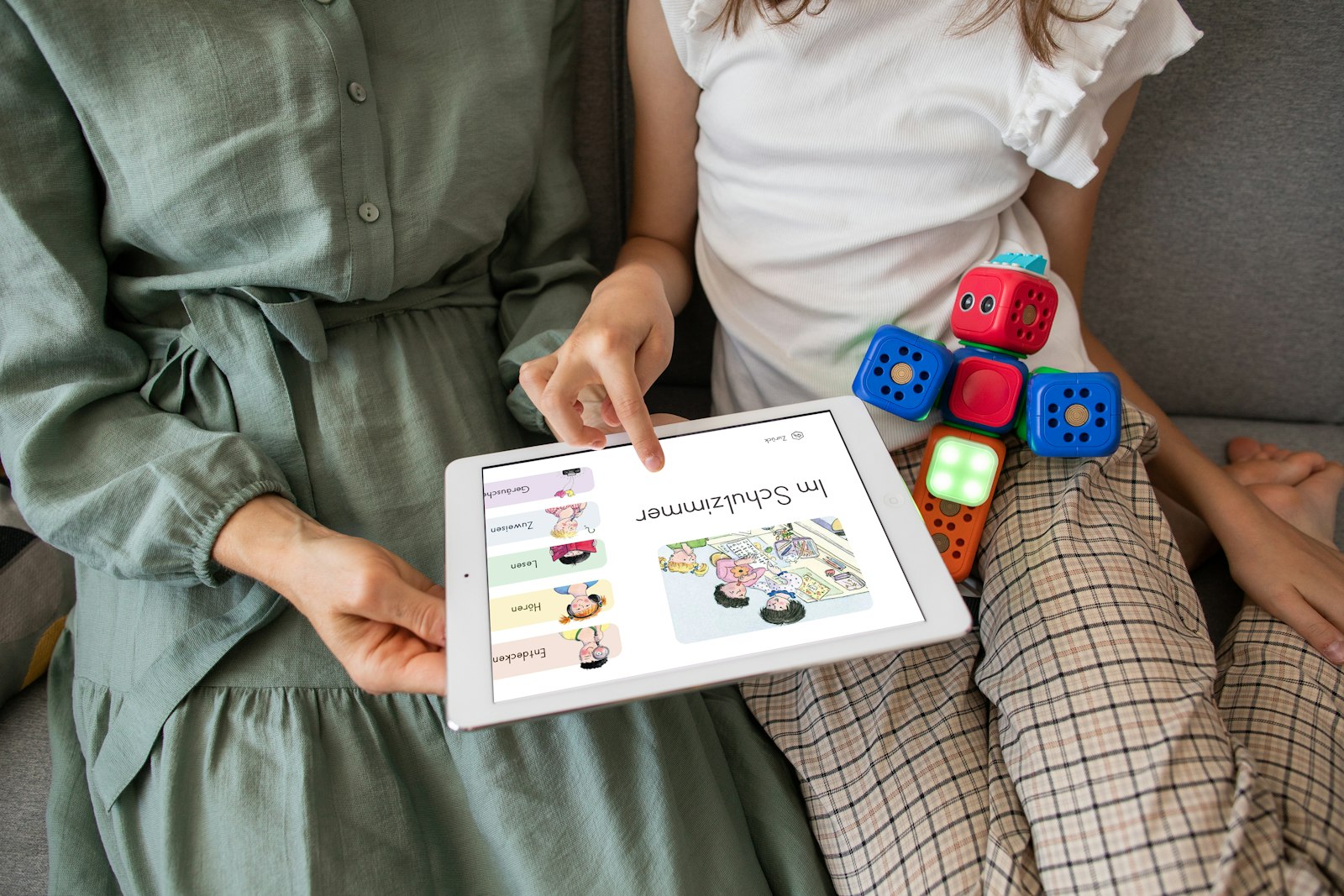 Two people sitting on a couch looking at a tablet displaying children's educational content. A toy with colorful cubes is on the child's lap.