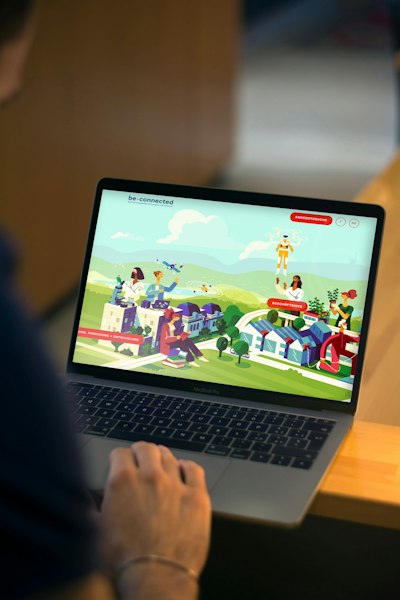 Person using a laptop displaying a webpage with colorful illustrations of people, buildings, and nature, featuring "be-connected" at the top.