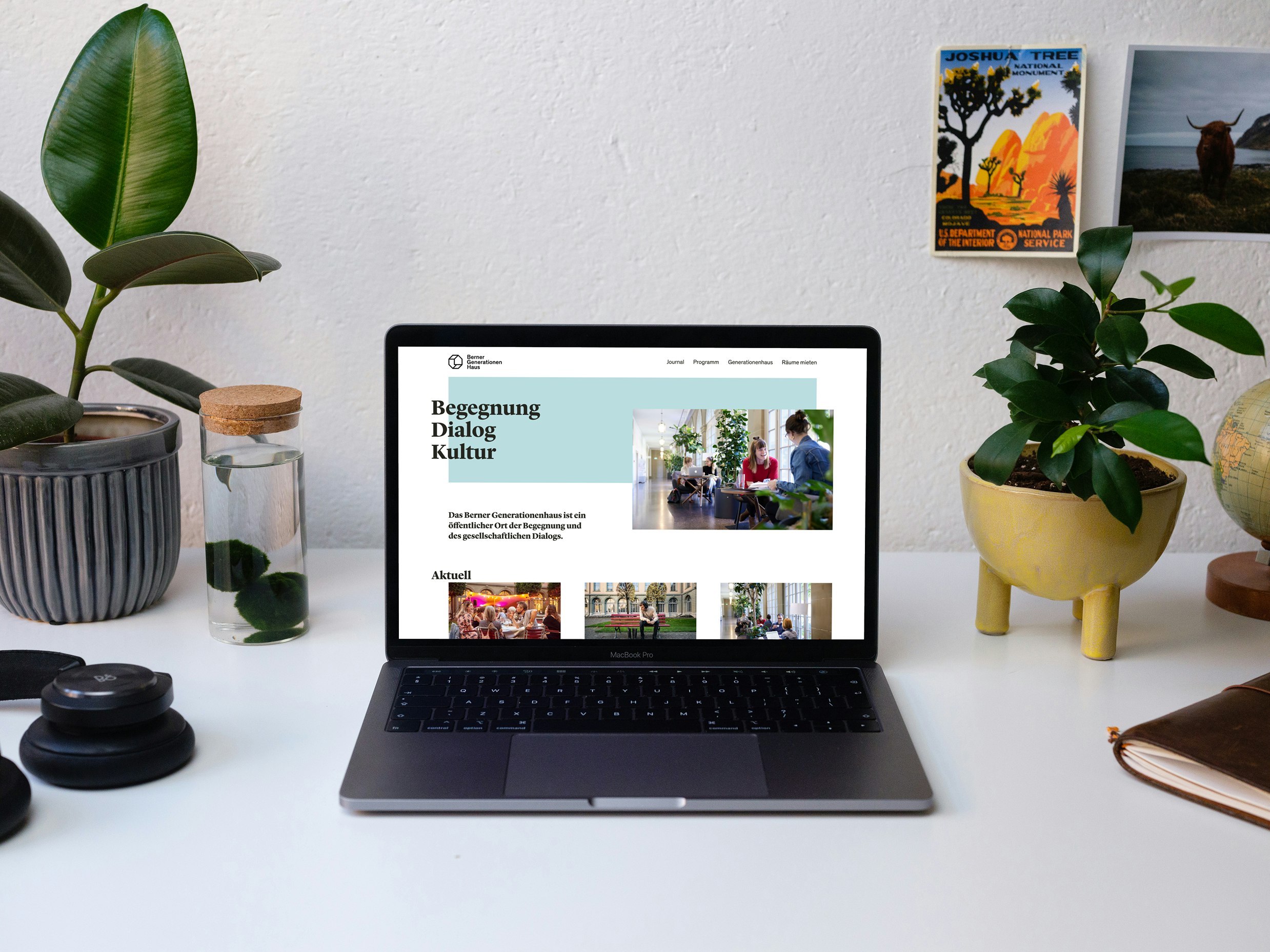 A laptop displaying a webpage on a desk with plants, a water bottle, and postcards in the background.
